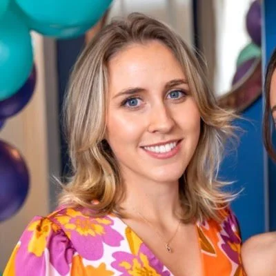 Smiling woman with blonde hair in a colorful floral dress, standing near purple and blue balloons in an indoor setting.