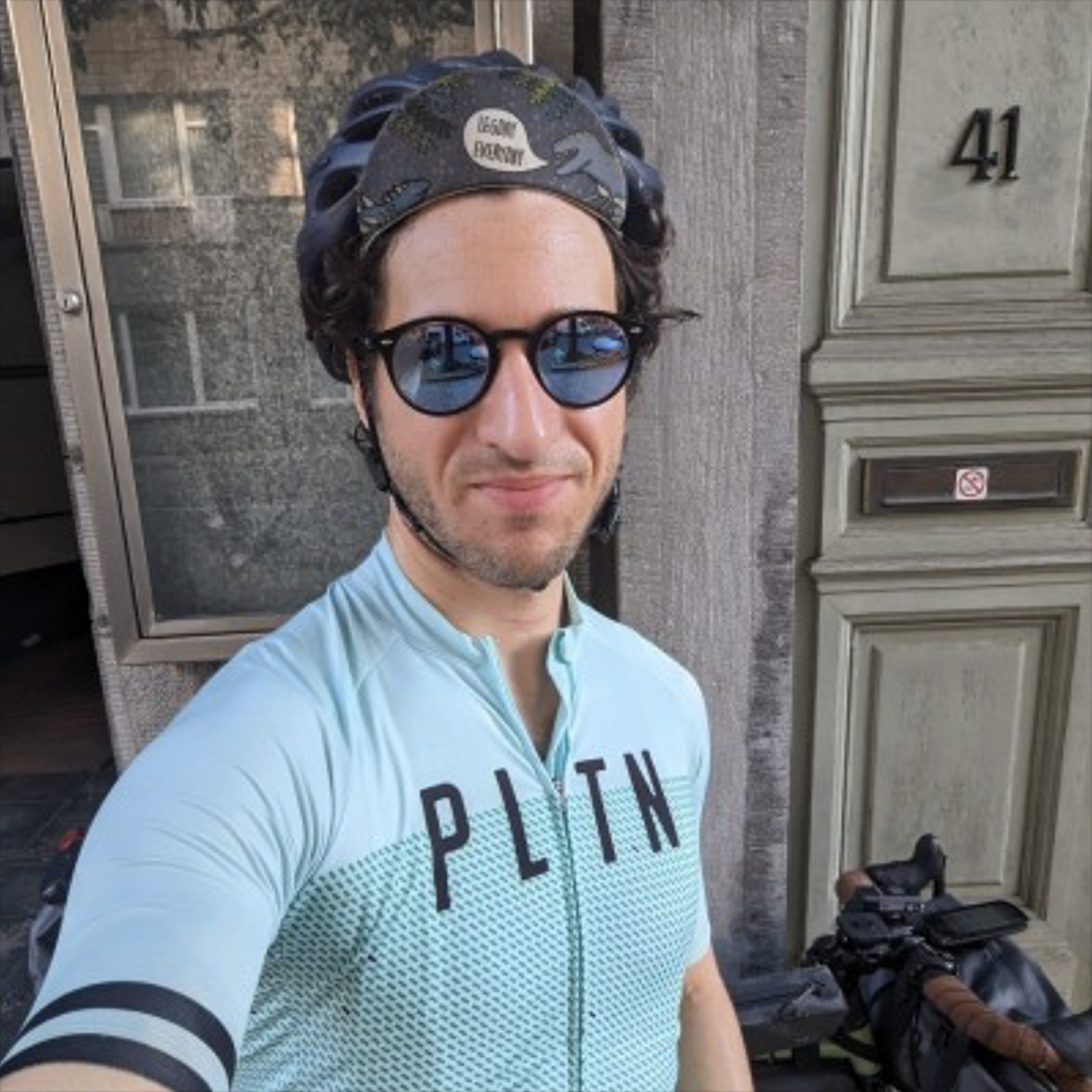 A man wearing cycling gear, including a light blue jersey with 'PLTN' printed on it, black sunglasses, a helmet, and a headband with a dinosaur design, standing outside in front of a building with the number 41 and a gray door, taking a selfie next to a bicycle.