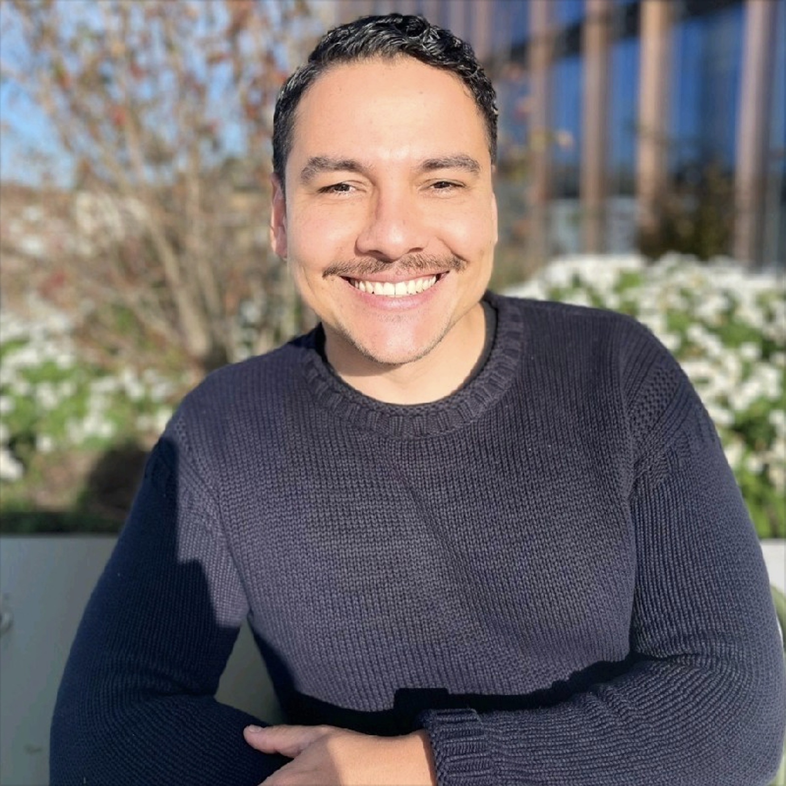 A smiling man with dark curly hair and a goatee, wearing a black knit sweater, sitting outdoors in front of a background of trees and buildings on a sunny day.