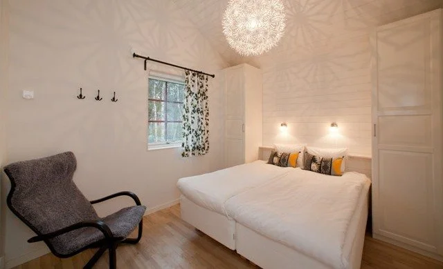 A bedroom with a bed, a gray armchair, a window with floral curtains, a white wardrobe, decorative pillows, and a modern chandelier ceiling light.