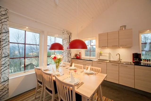 Dining area with a wooden table set with plates, glasses, and silverware, surrounded by wooden chairs, in a bright kitchen with large windows, red pendant lights, and white cabinetry.
