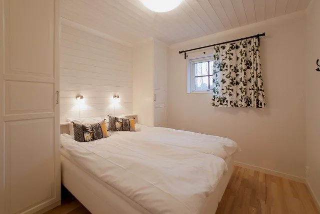 A cozy bedroom with a large bed, white bedding, and decorative pillows. White walls, wood flooring, a window with floral curtains, and built-in closets.