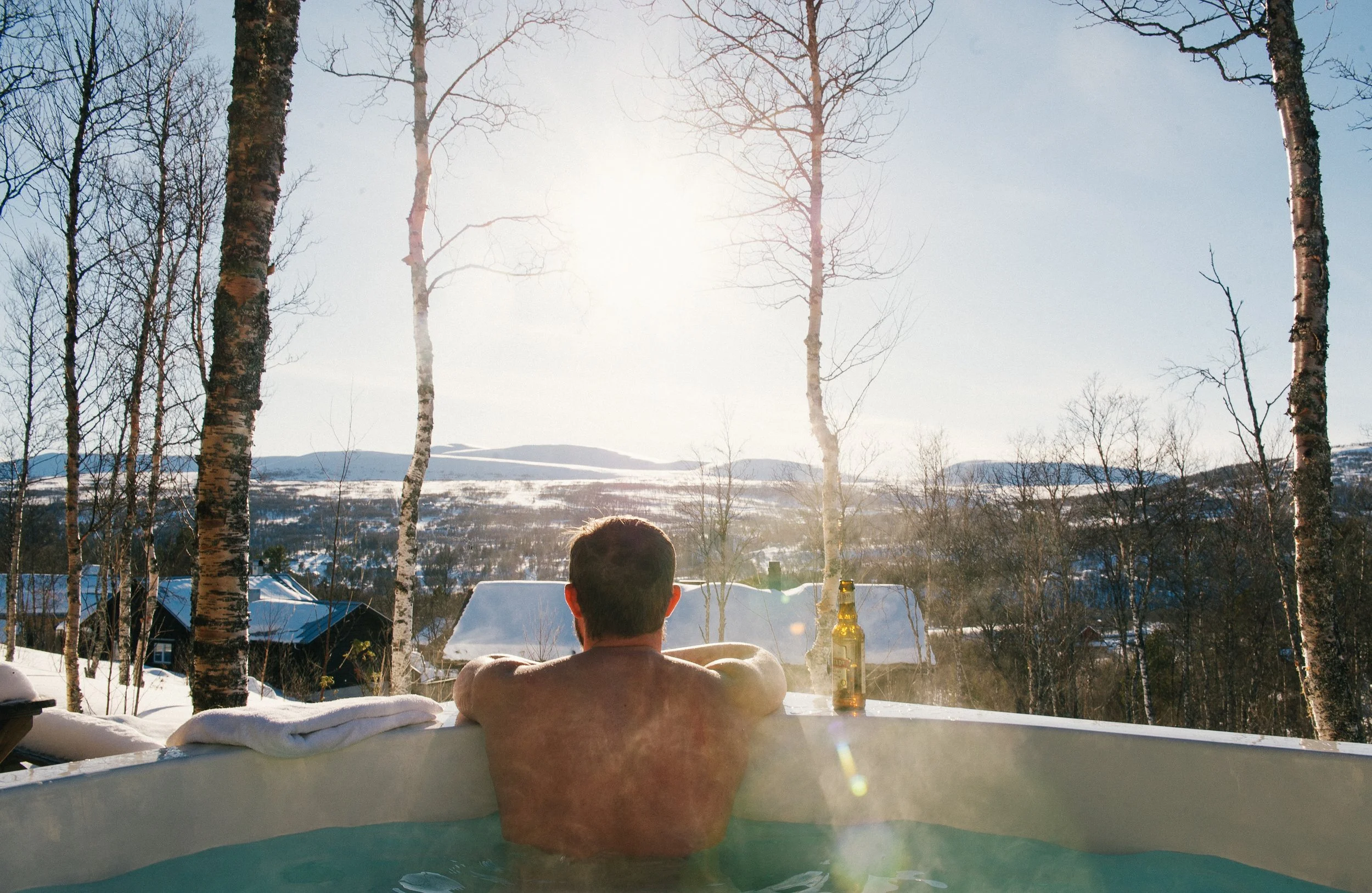 A man relaxing in an outdoor hot tub during winter, facing a snowy landscape with bare trees, snow-covered rooftops, and mountains in the distance, with the sun low in the sky.