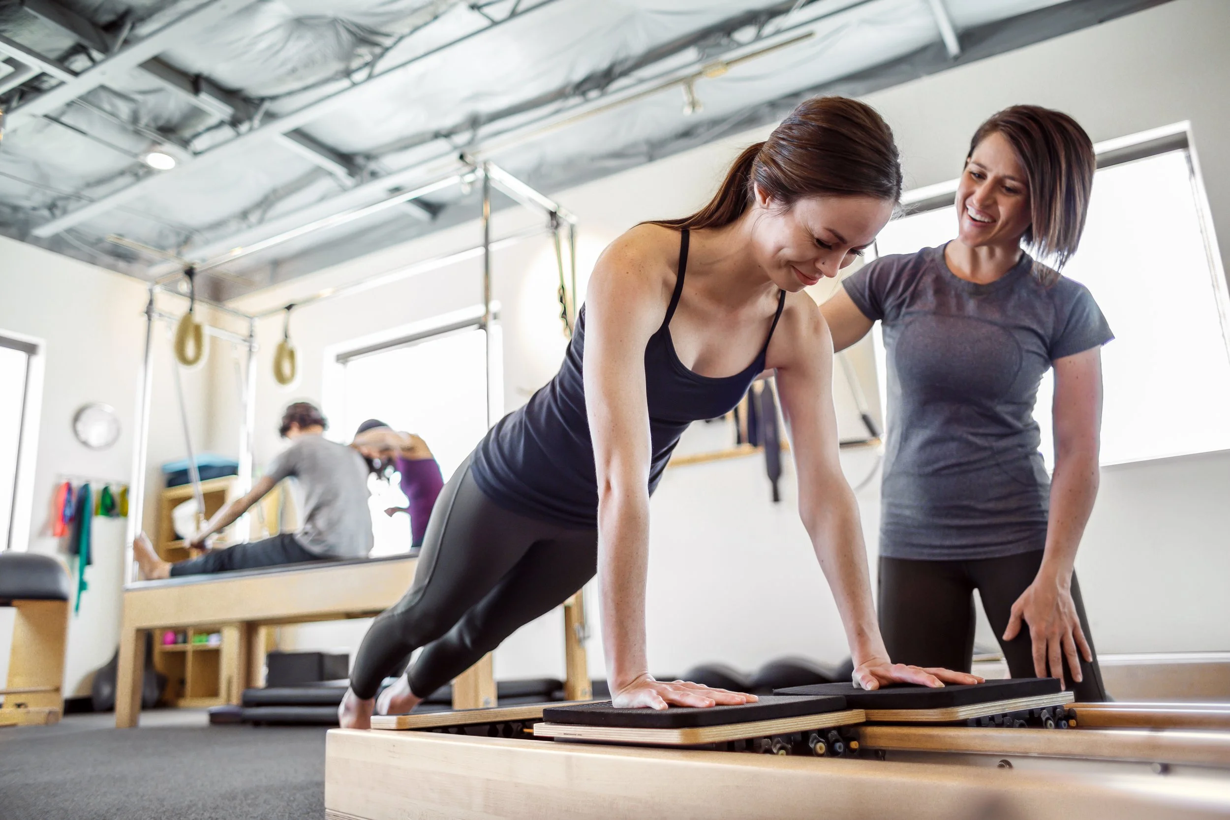 A woman doing a plank exercise on a reformer machine in a gym, supervised by a pilates trainer, with other people exercising in the background.