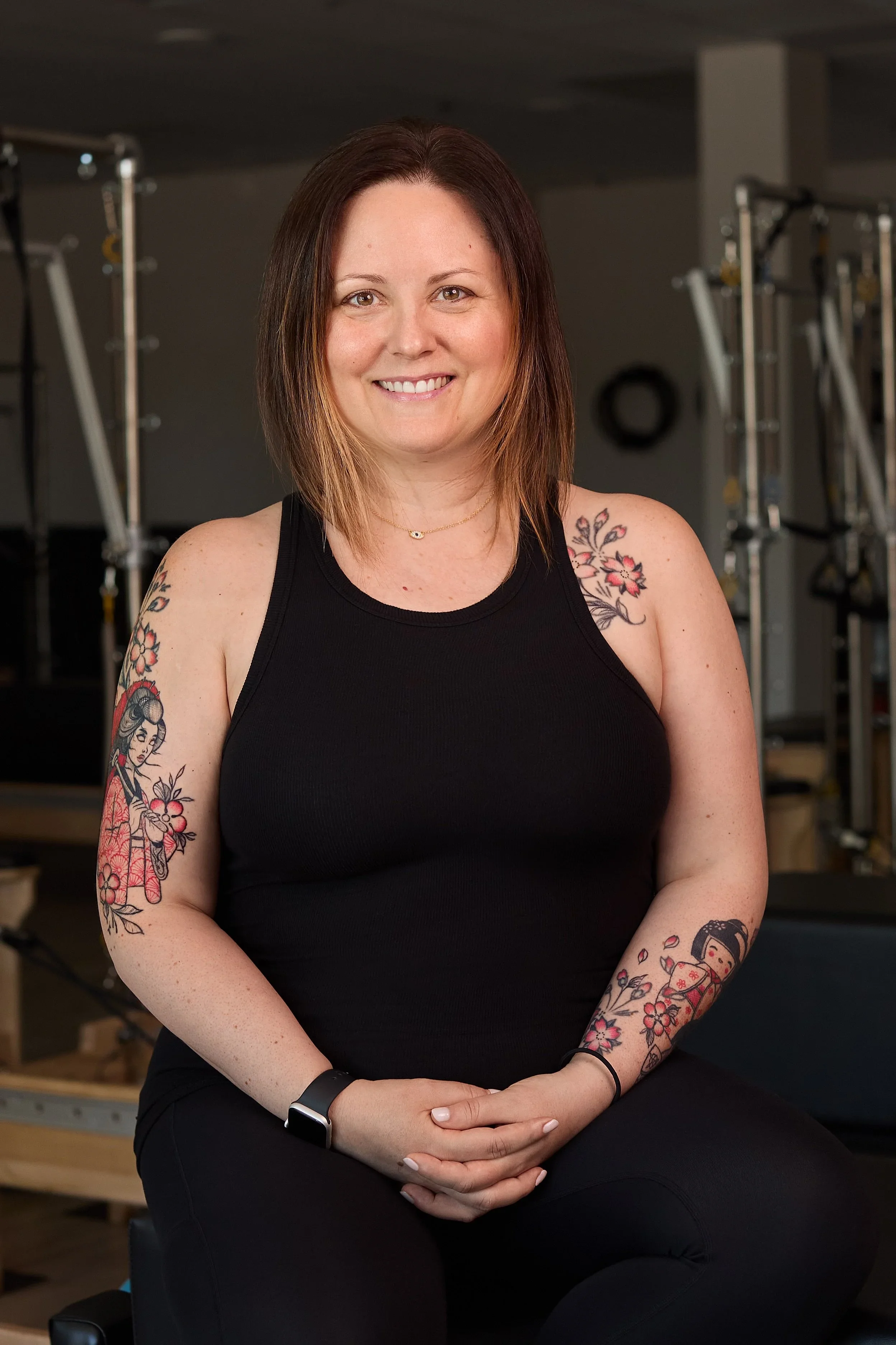 A woman with shoulder-length brown hair and tattoos on her arms, wearing a black tank top, sitting and smiling at the camera in a gym setting.