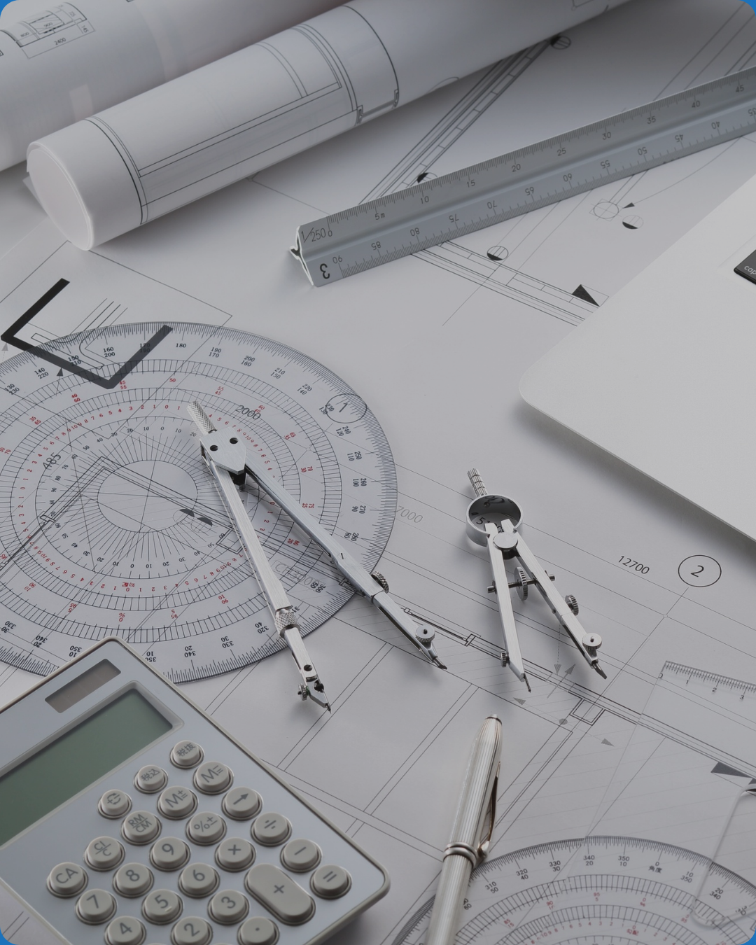 Blueprints, drafting tools including a protractor, compasses, rulers, a calculator, and a pen on a desk, indicating architectural or engineering work.