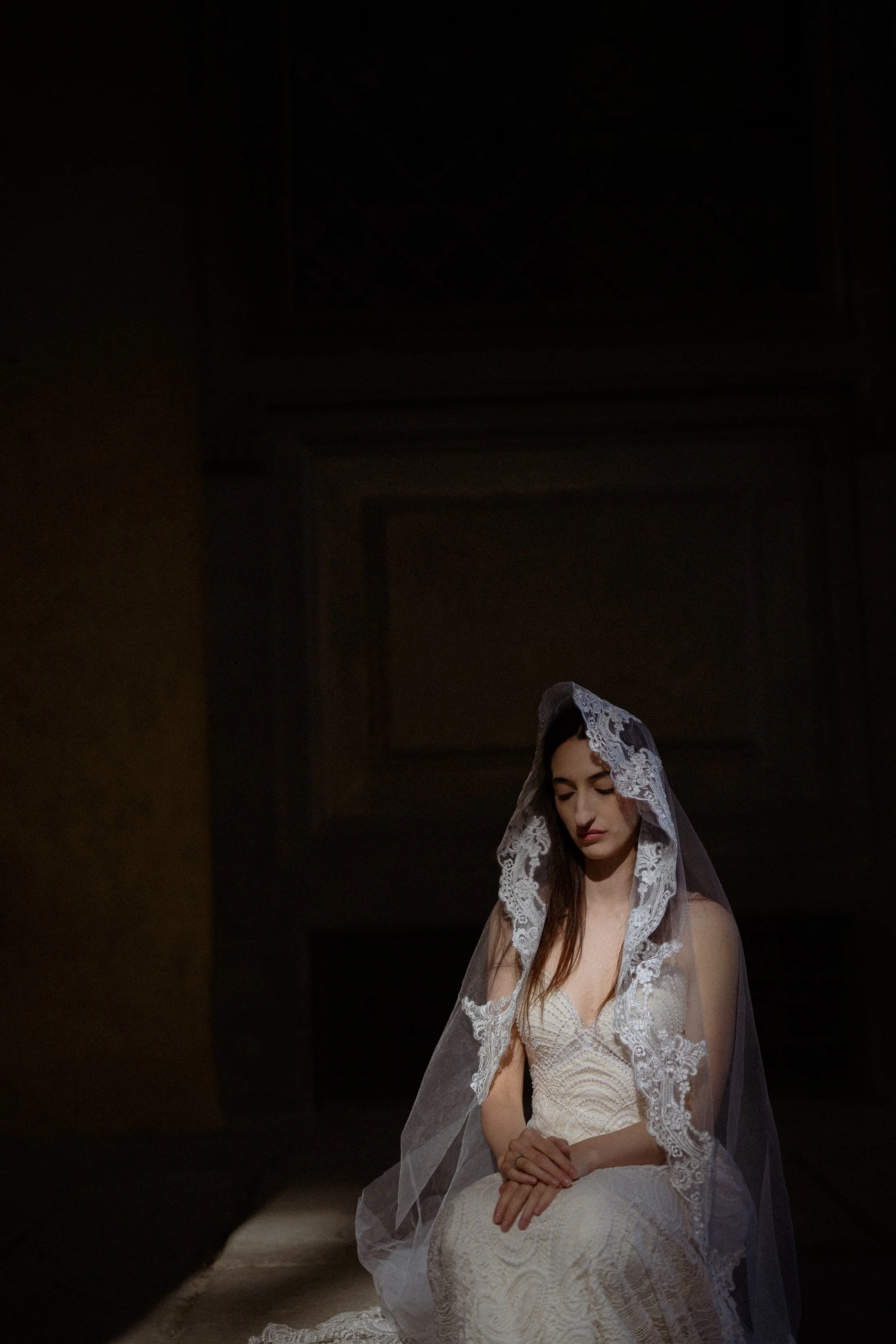 A woman in a wedding dress and lace veil sitting with her hands clasped, in a dimly lit room with a dark background.