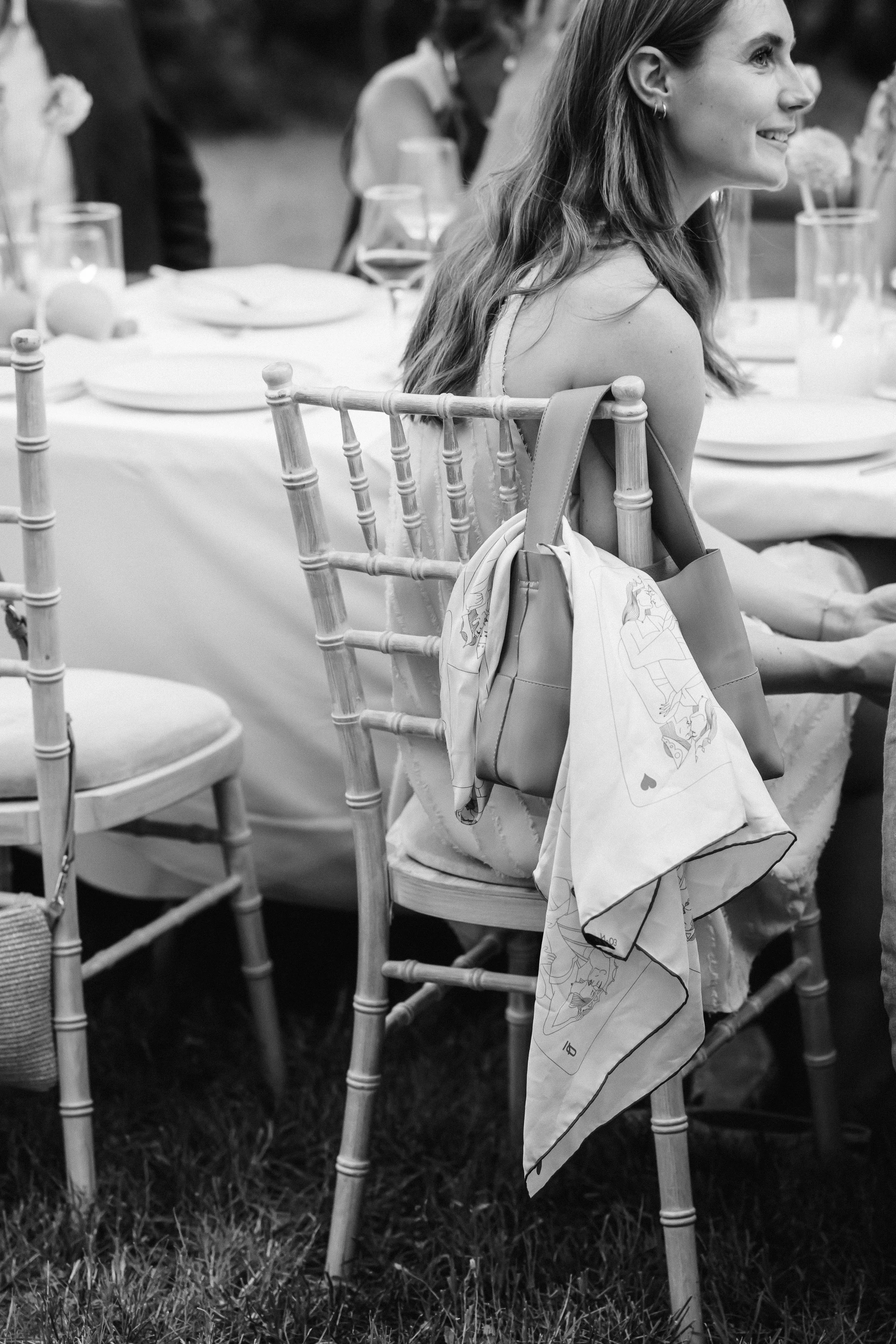 A woman with long hair smiling at a formal outdoor event, sitting on a wooden chair with a large handbag and a scarf hanging from it. The table is decorated with glasses, plates, and floral centerpieces.