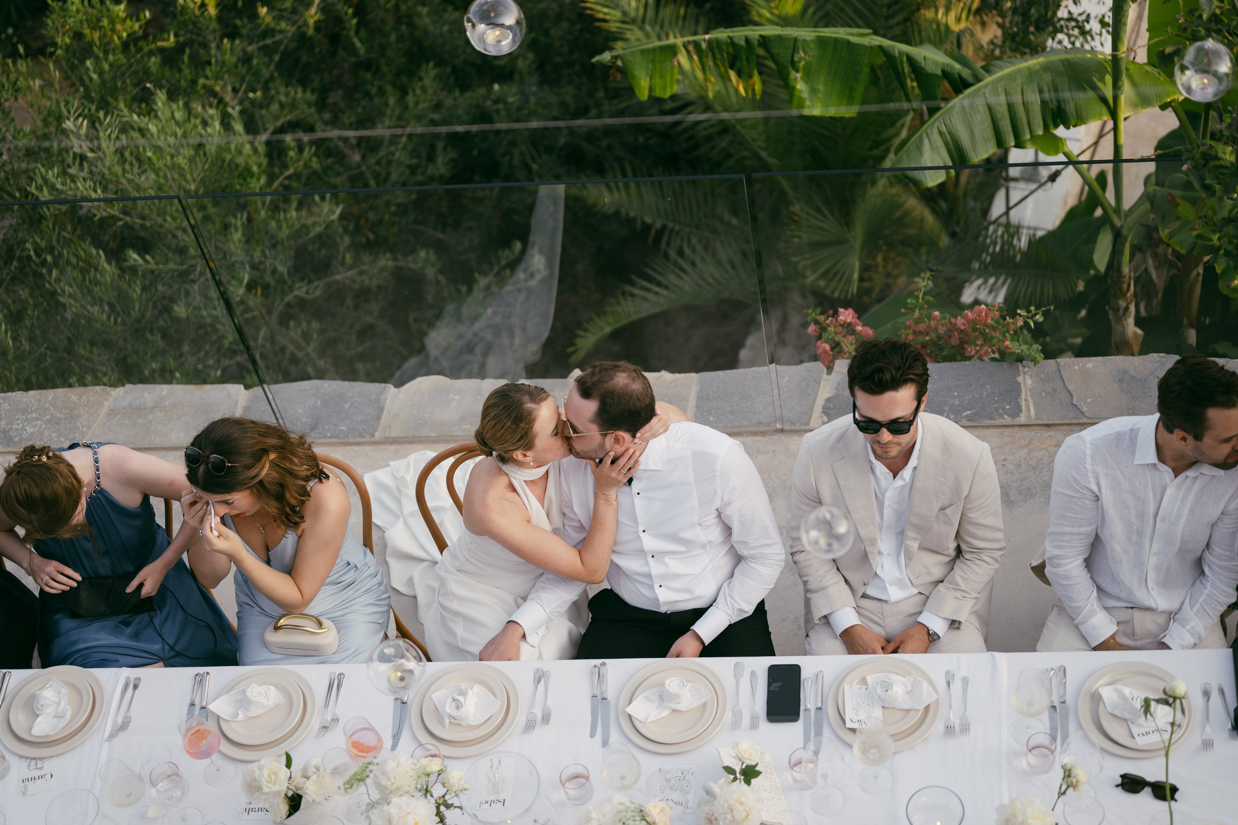 People sitting at a long reception table, with a couple kissing in the center, during an outdoor event or wedding reception.