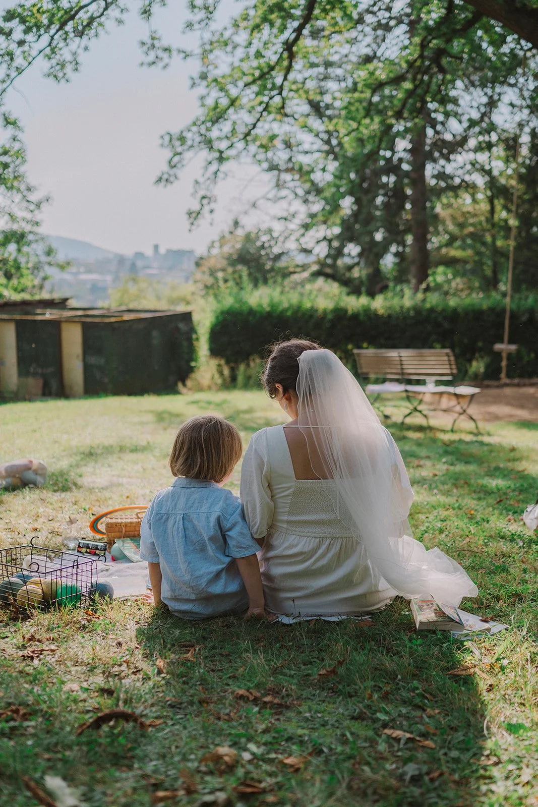 A woman dressed as a bride with a veil sitting on grass with a young boy, probably a child, in a park or garden area with trees, benches, and a cityscape in the background.