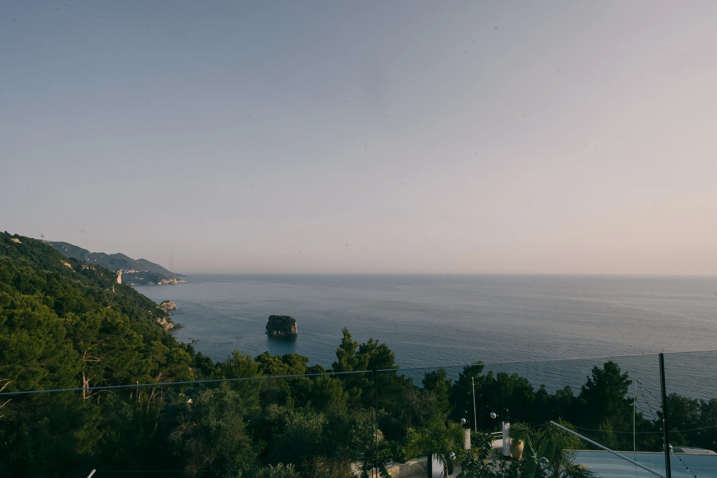 Scenic coastal view with calm ocean, rocky cliffs, and lush green trees under a clear sky.