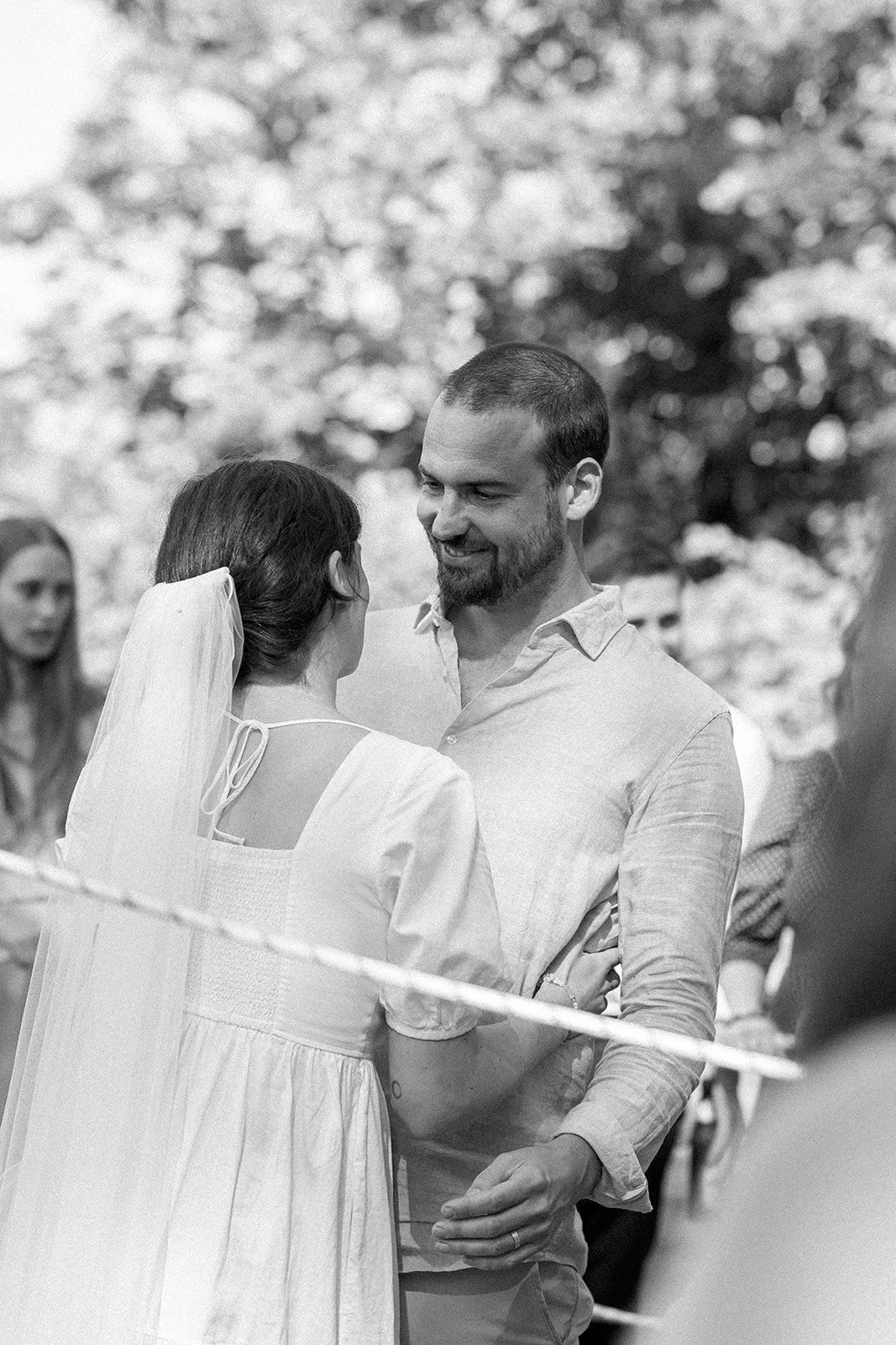 A black-and-white photo of a wedding ceremony, showing a couple sharing a dance or intimate moment, with guests in the background.