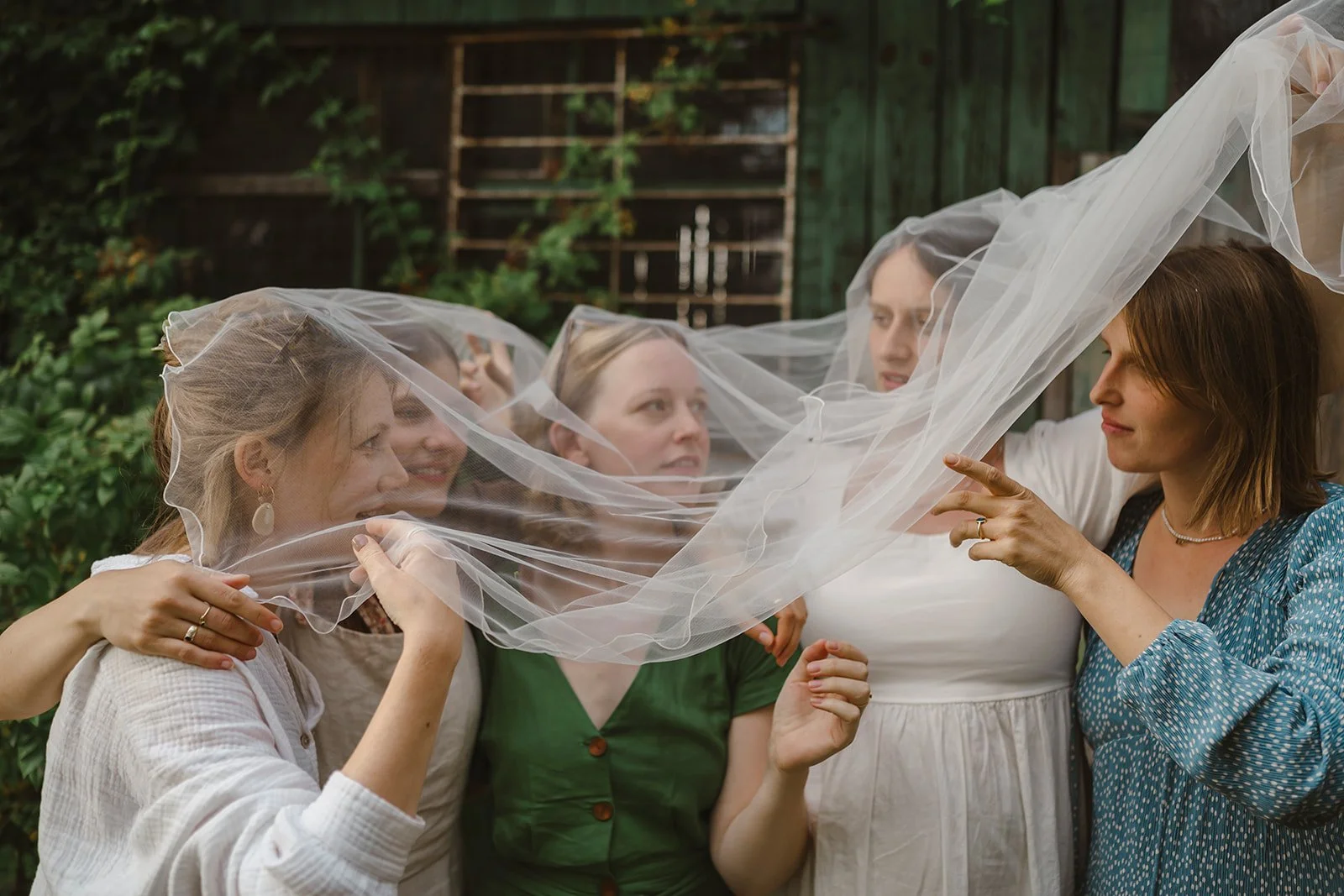 Five women gathered outdoors, holding a sheer veil between them, in a garden setting with greenery and a wooden fence in the background.