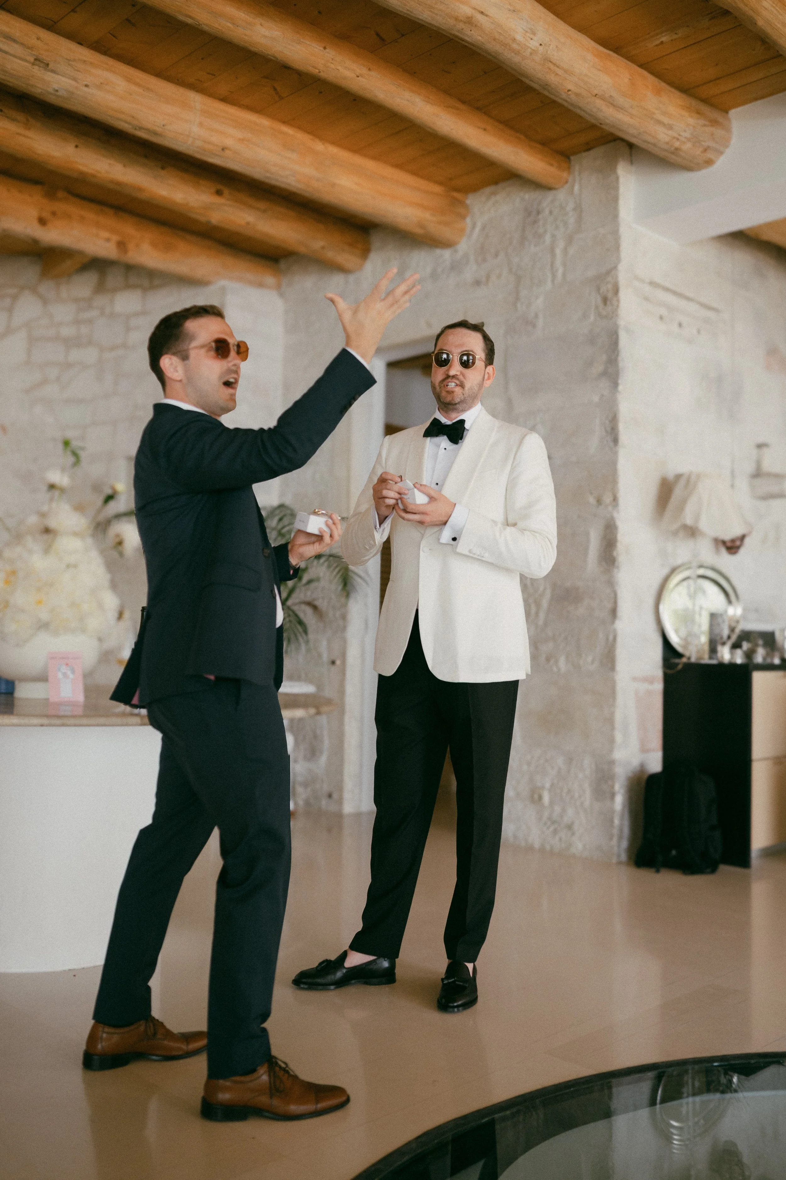 Two men wearing suits and sunglasses are having a conversation indoors, with one gesturing animatedly while the other listens. The setting appears to be a modern, rustic room with exposed wooden beams and stone walls, decorated with flowers and artwo