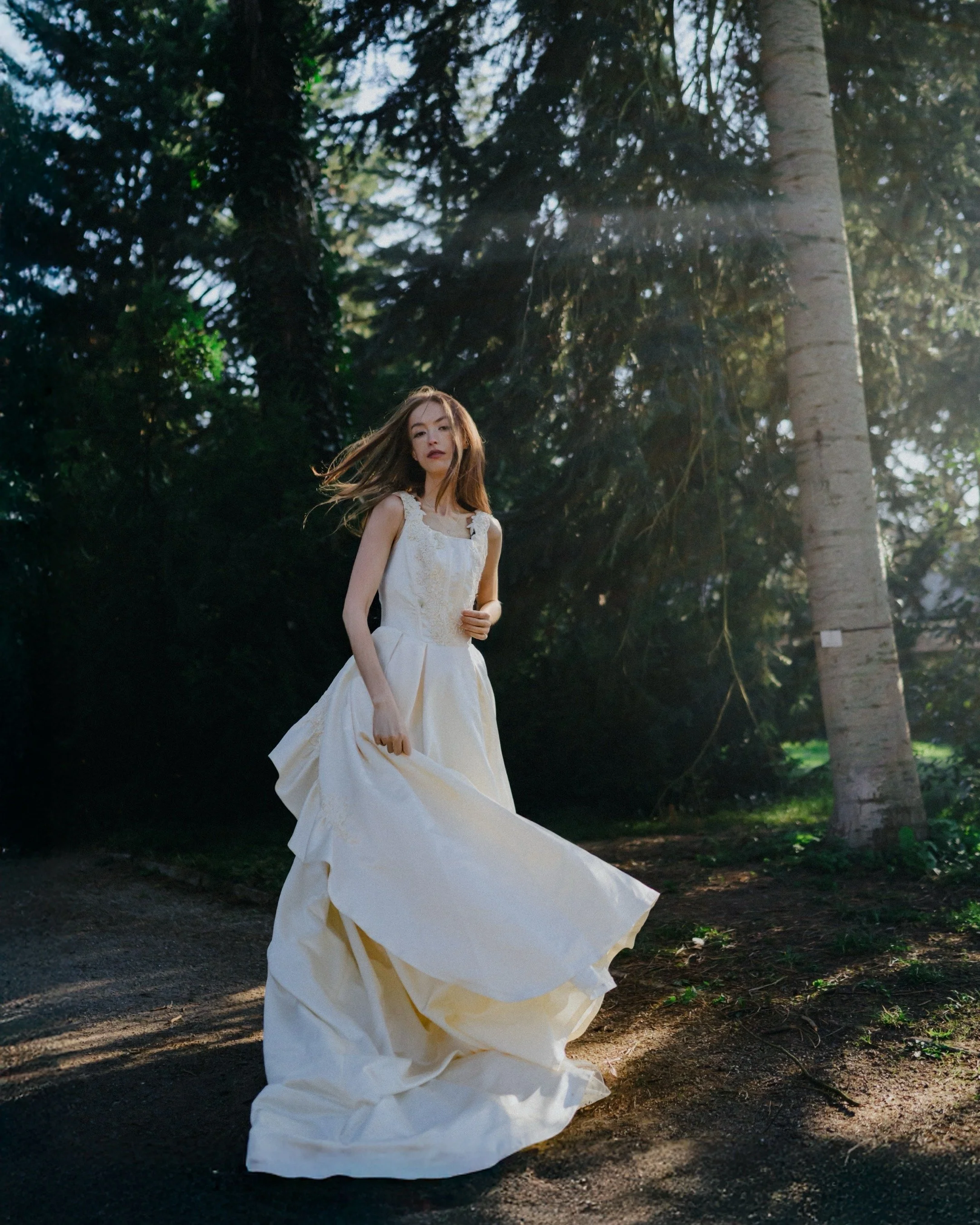 A woman in a white wedding dress standing on a forest path surrounded by trees, with sunlight shining through the branches.