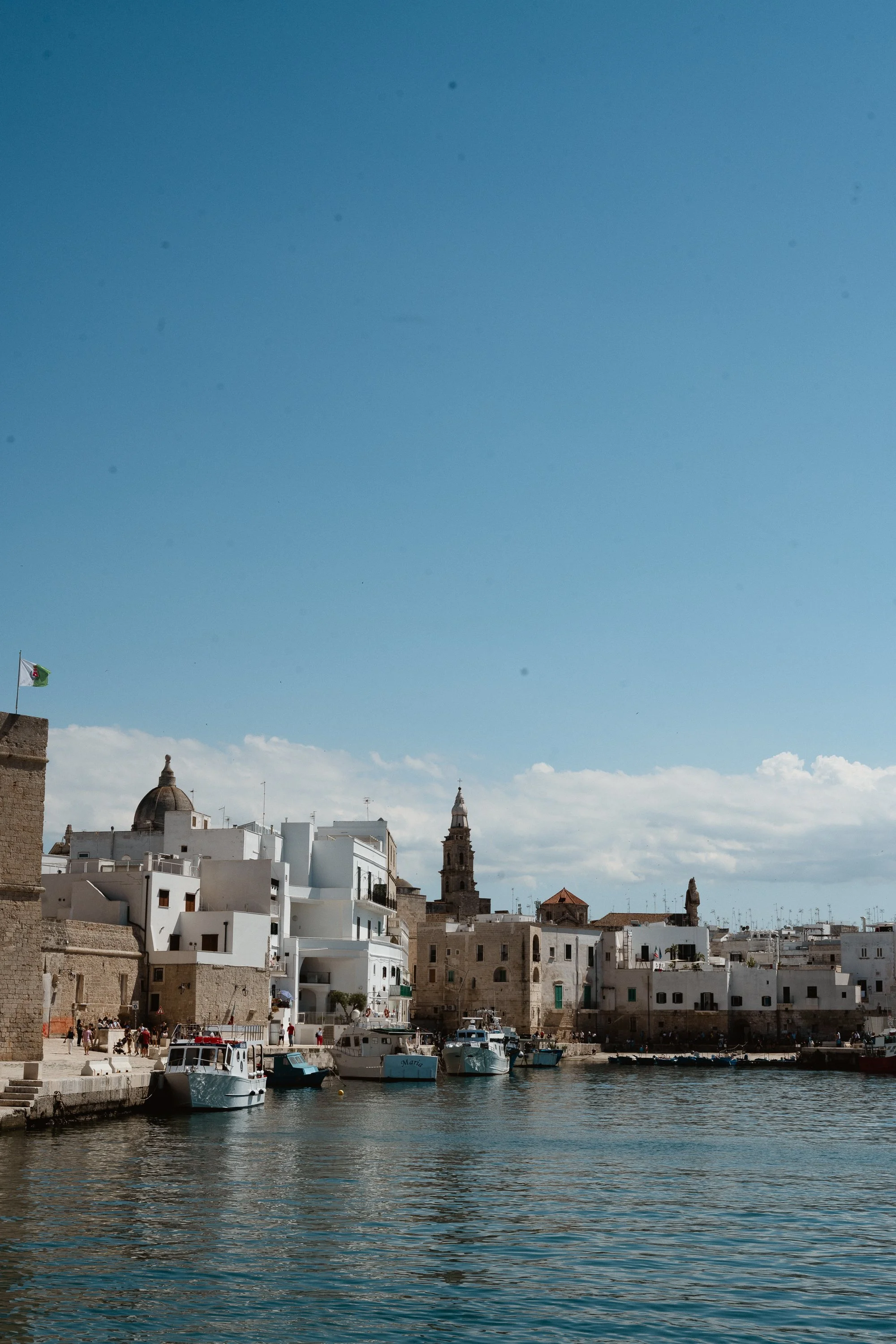 A harbor with boats docked along the pier in front of historic white buildings and towers under a clear blue sky.