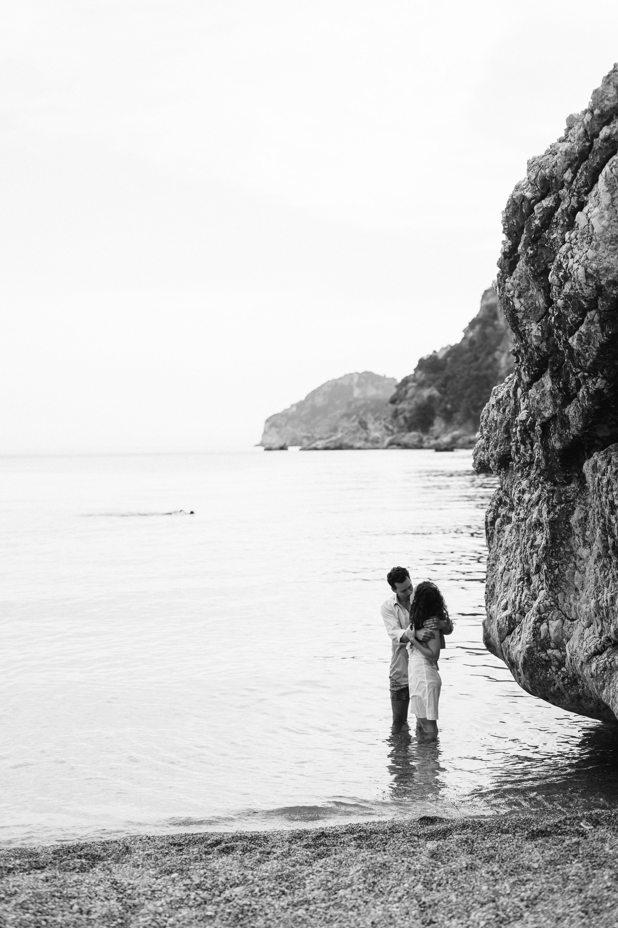 A black and white photo of a couple holding each other in shallow water by a rocky shore on a beach, with cliffs in the distance.