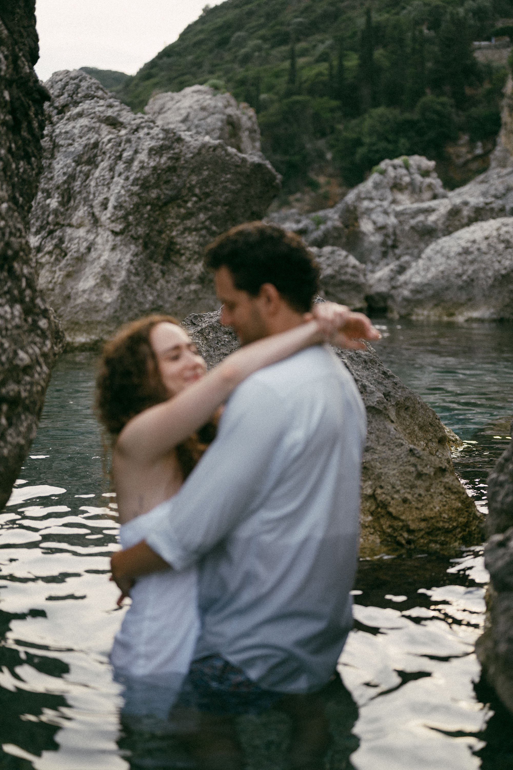 A couple embraces in a rocky river surrounded by nature with green hills in the background.