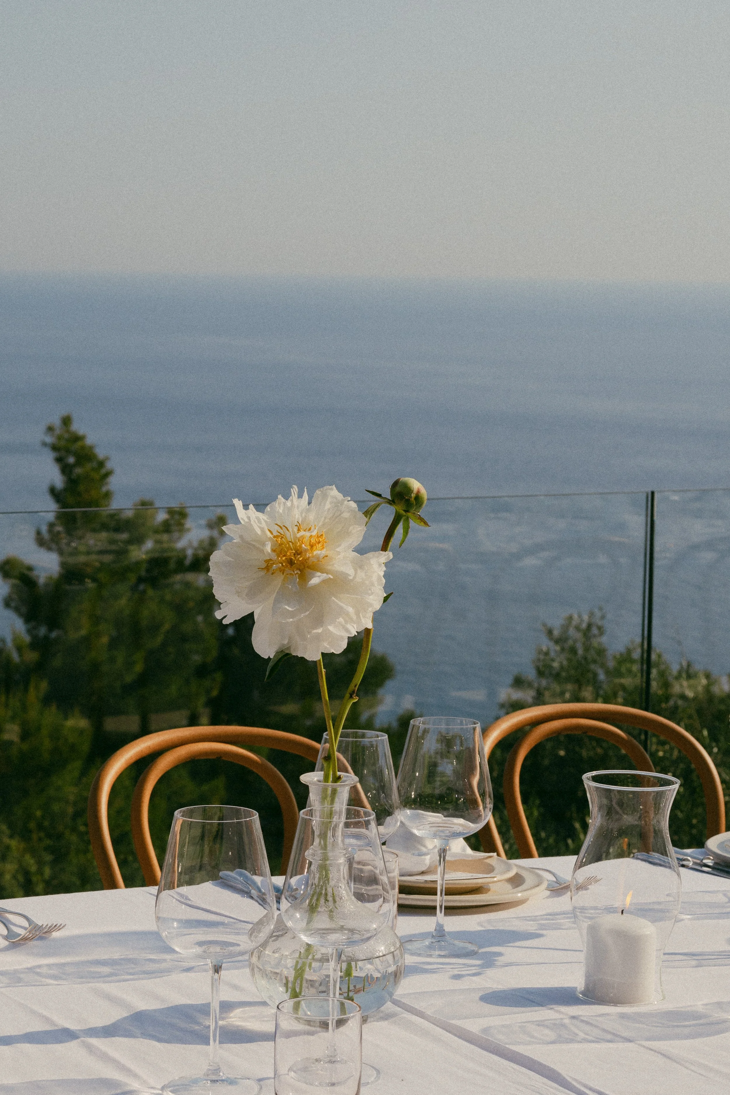 Elegant outdoor dining table with a white tablecloth, glassware, a candle, and a vase with a white peony flower, overlooking a scenic ocean view with trees.