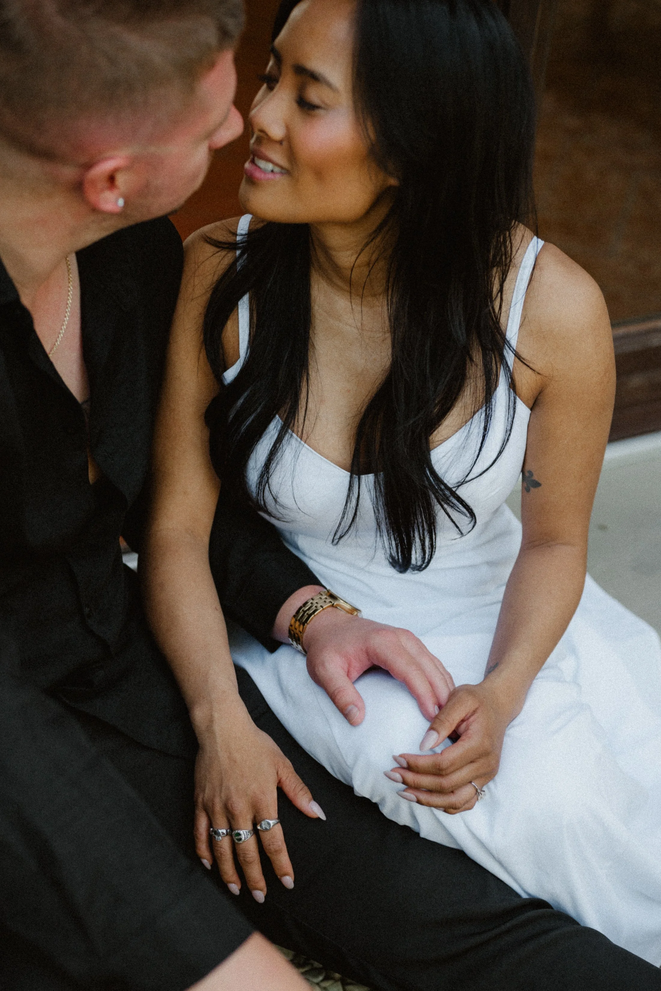 A man and woman sitting very close, touching, looking at each other affectionately, with the woman wearing a white dress and the man in a black shirt, sharing an intimate moment.