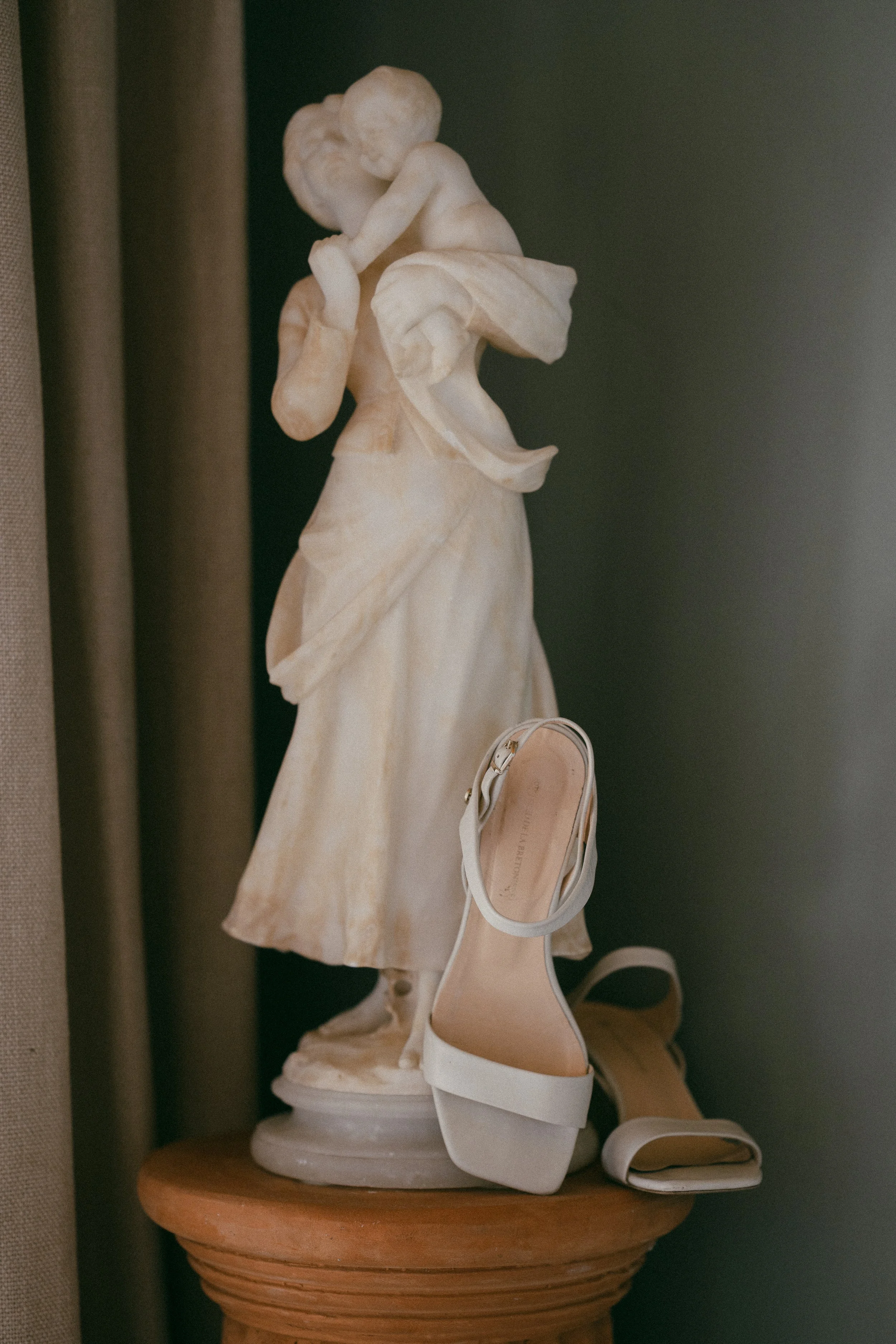 A white marble angel sculpture on a round wooden pedestal, with a pair of white high heel shoes placed next to it. The background includes neutral-colored curtains and a dark wall.