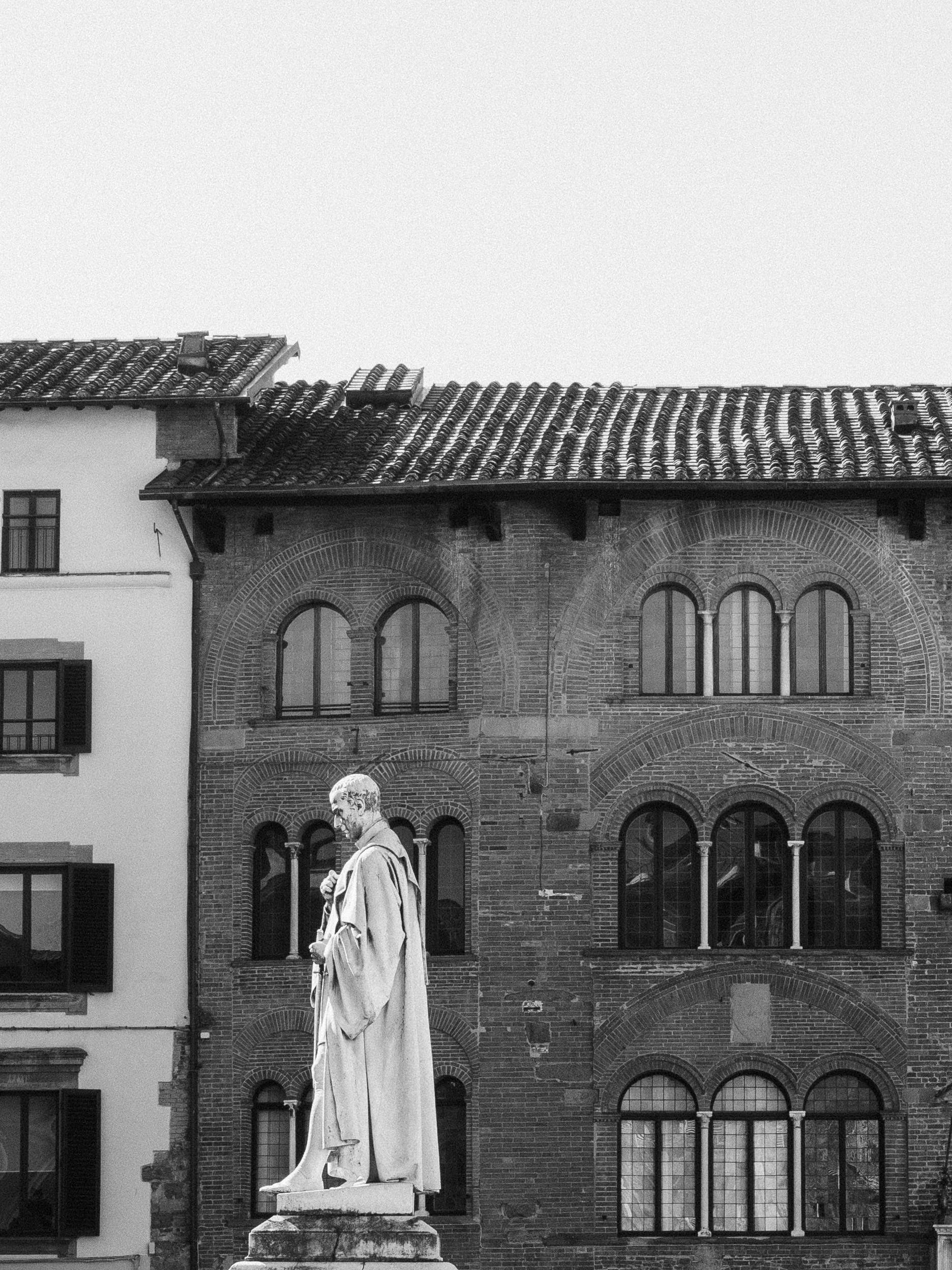 A black and white photo of a statue of a man in a long robe standing on a pedestal, with a historic brick building with arched windows in the background.