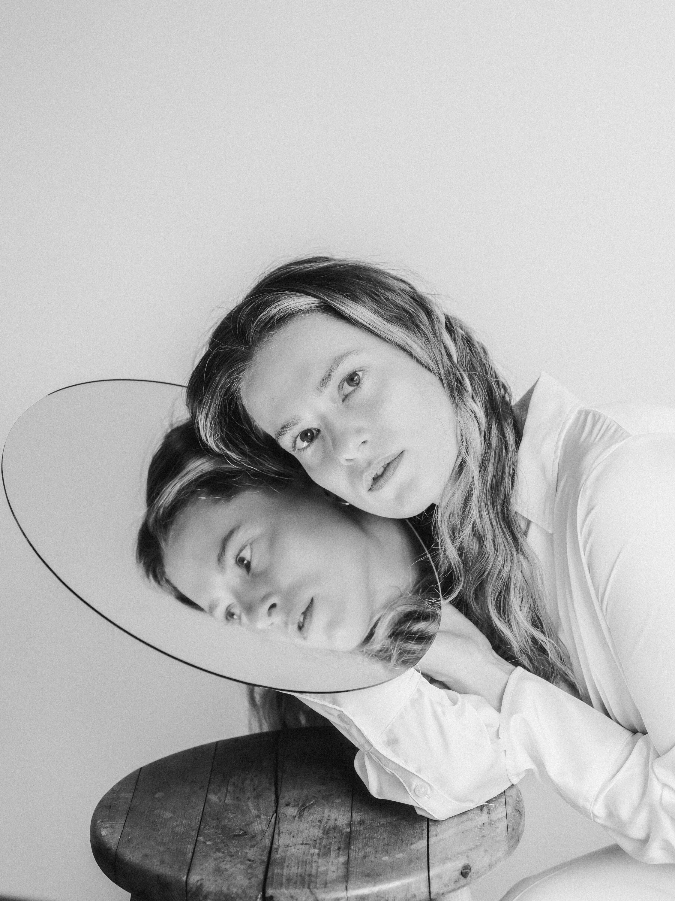 A woman wearing a white shirt and oversized hat, resting her head with her cheek against a mirror that reflects her face, showing her head tilted to the side and her eyes open, on a wooden stool in a minimal setting.