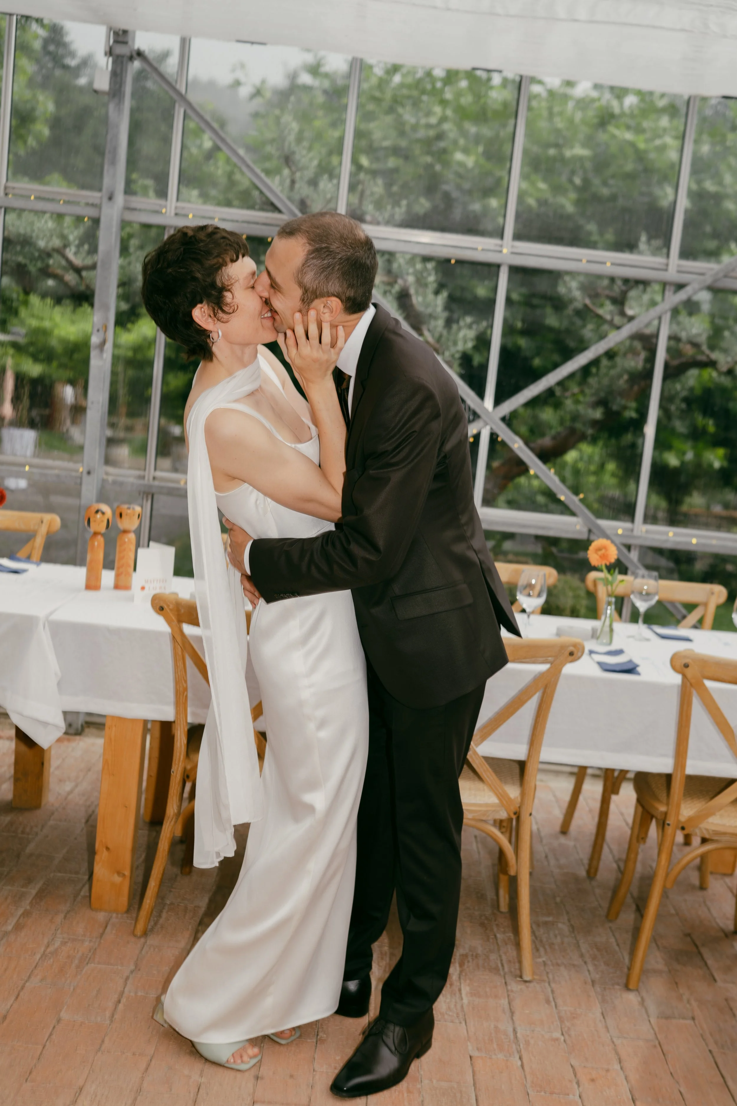 A couple is kissing, with the woman holding the man's face, in an indoor setting with large windows, trees visible outside, wooden chairs, and a table decorated with flowers and wine glasses.