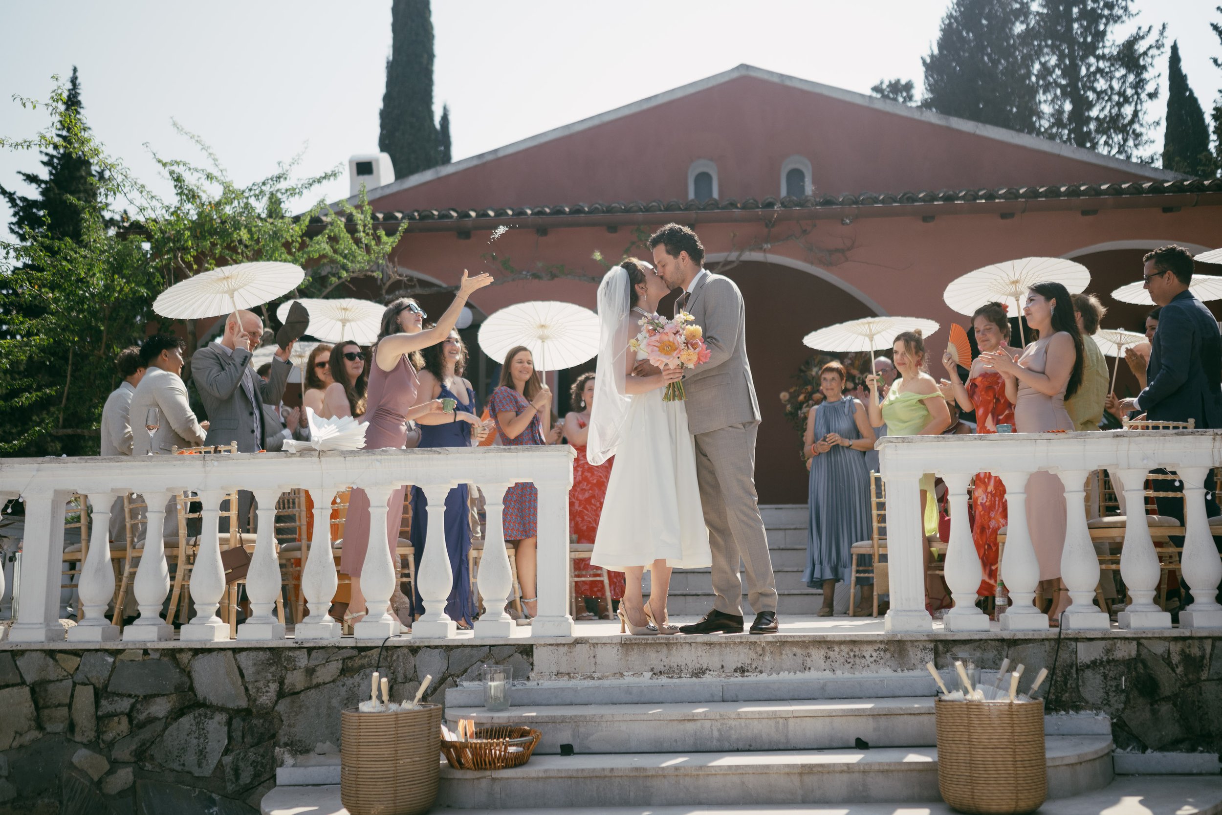 A wedding ceremony with a bride and groom kissing on a stone staircase, surrounded by guests holding parasols in an outdoor setting.