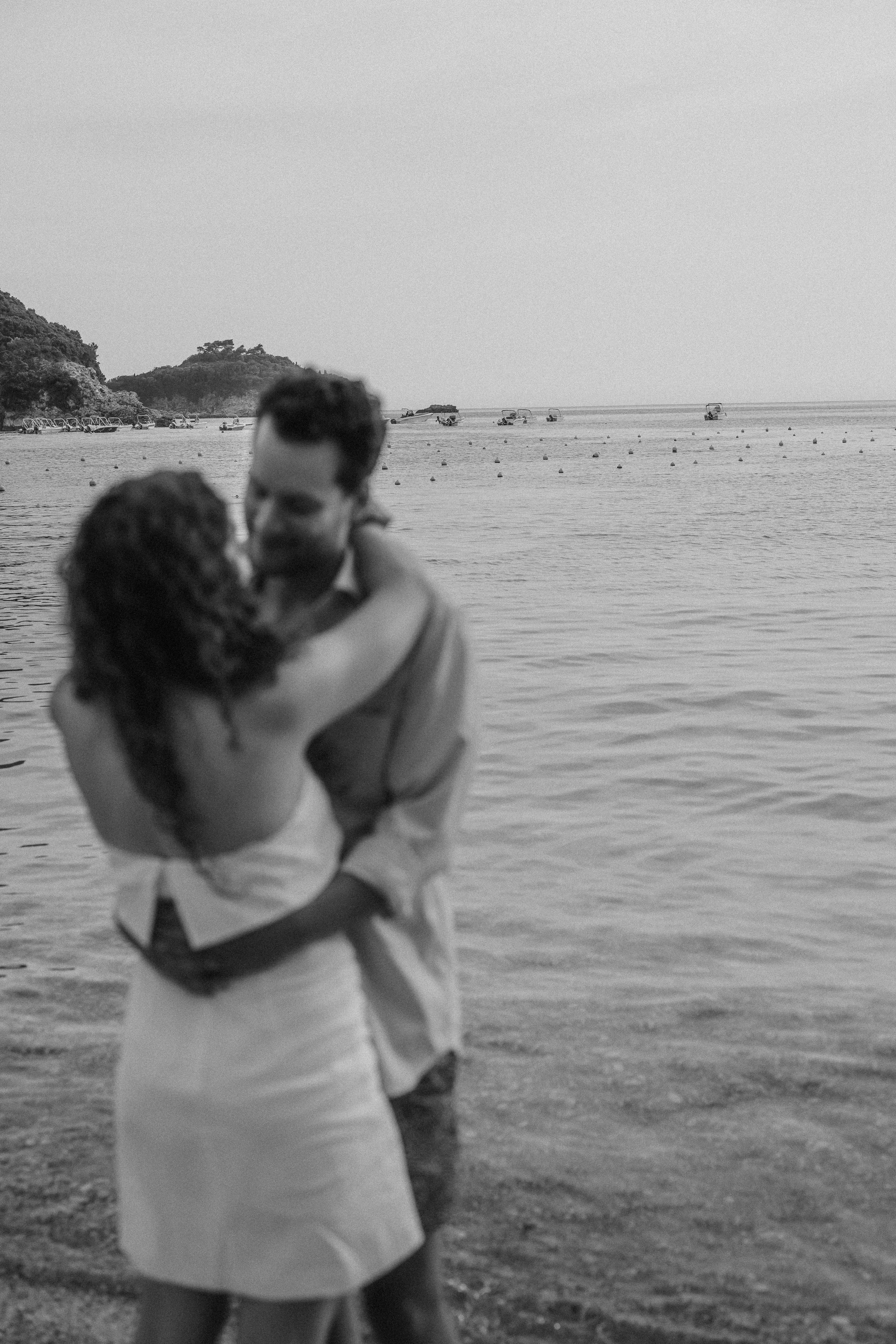 A couple embracing on a beach, with water and boats in the background.