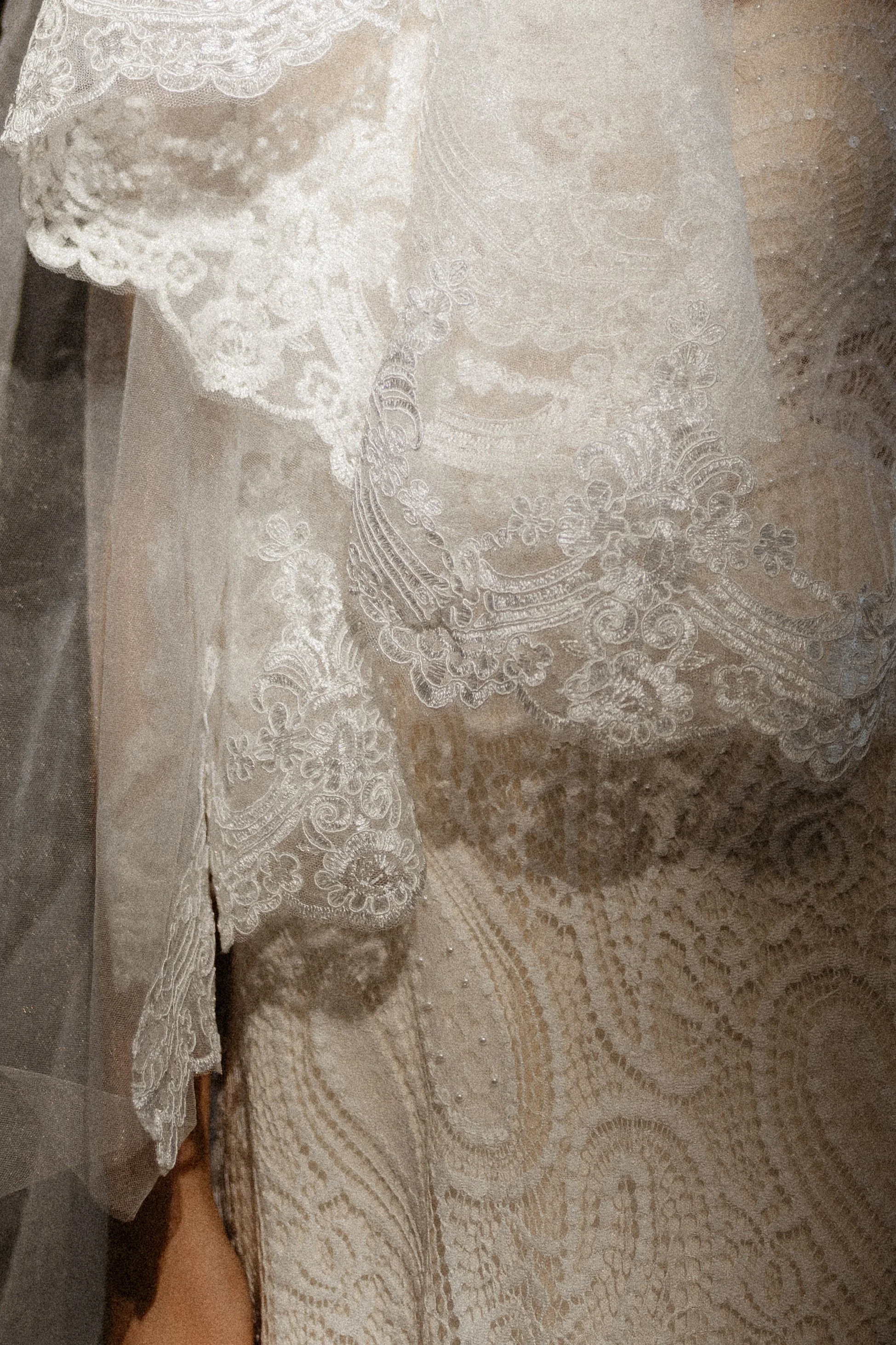 Close-up of intricate lace fabric, likely part of a wedding dress or formal gown, with detailed floral and swirl patterns.