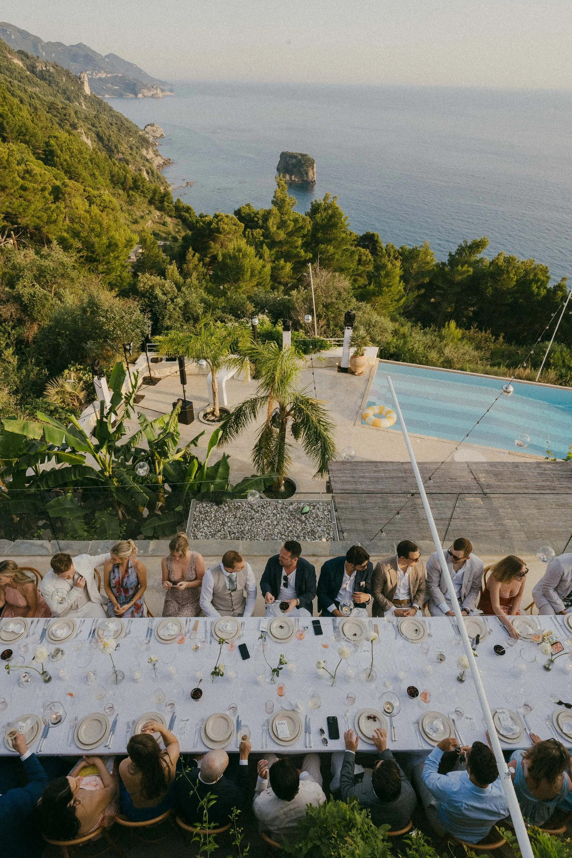 A long outdoor dining table with guests, set with plates, glasses, and floral centerpieces, overlooking a lush hillside and the ocean with a rocky island in the background, on a clear day.