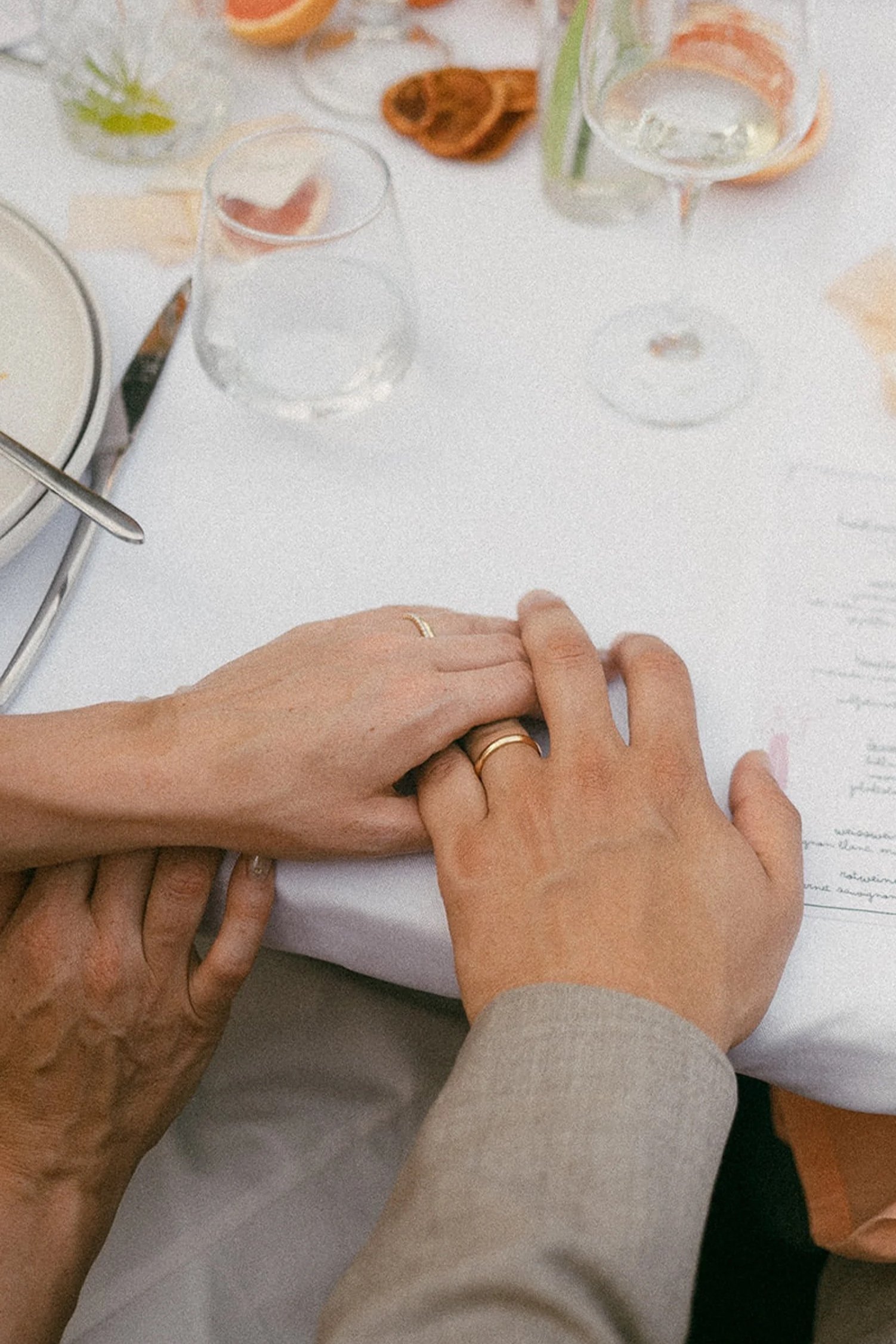 Two people holding hands on a white table with glasses of water, dishes, and a menu.