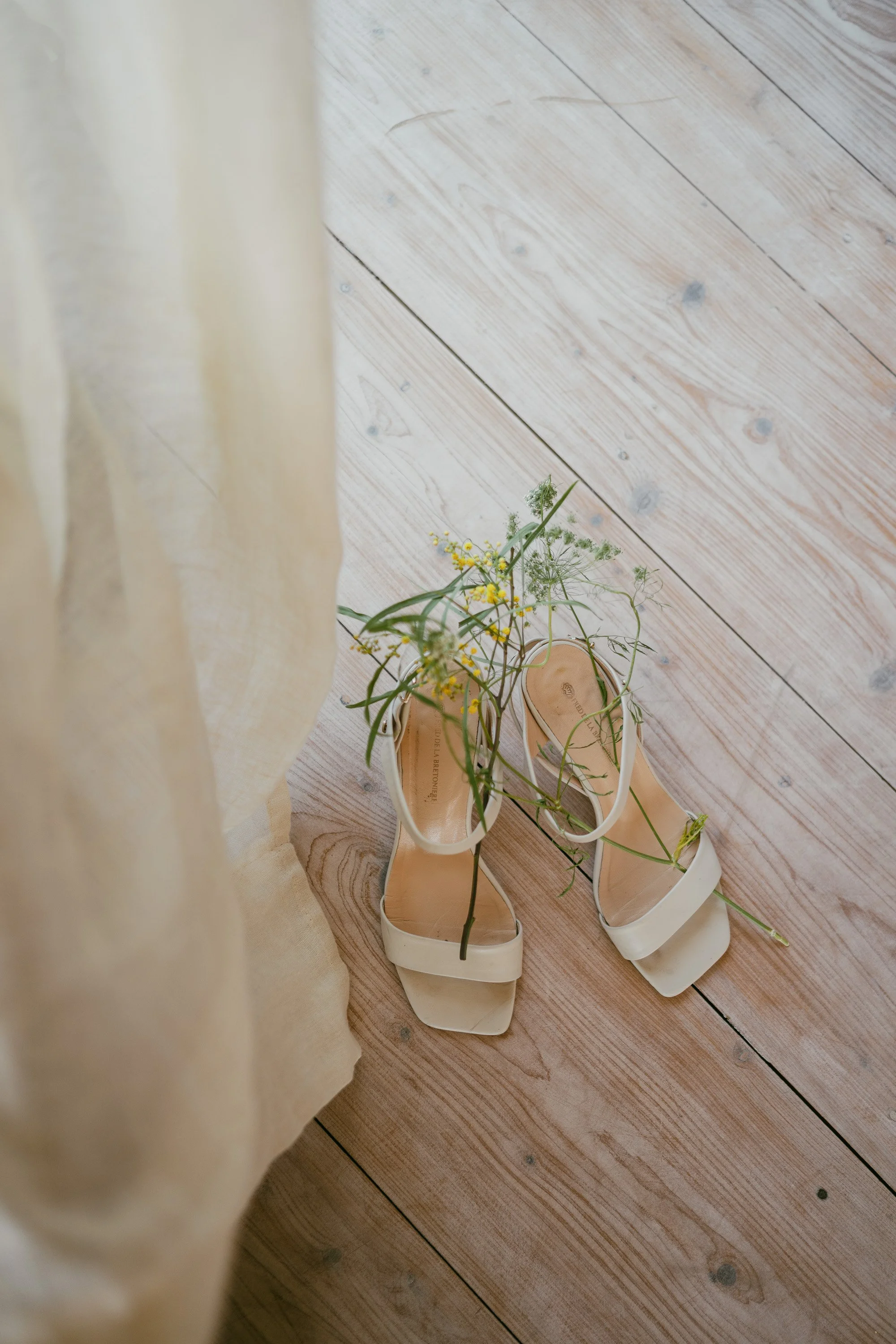 White high-heeled shoes with beige flowers and green foliage inside, on a wooden floor near a cream curtain.