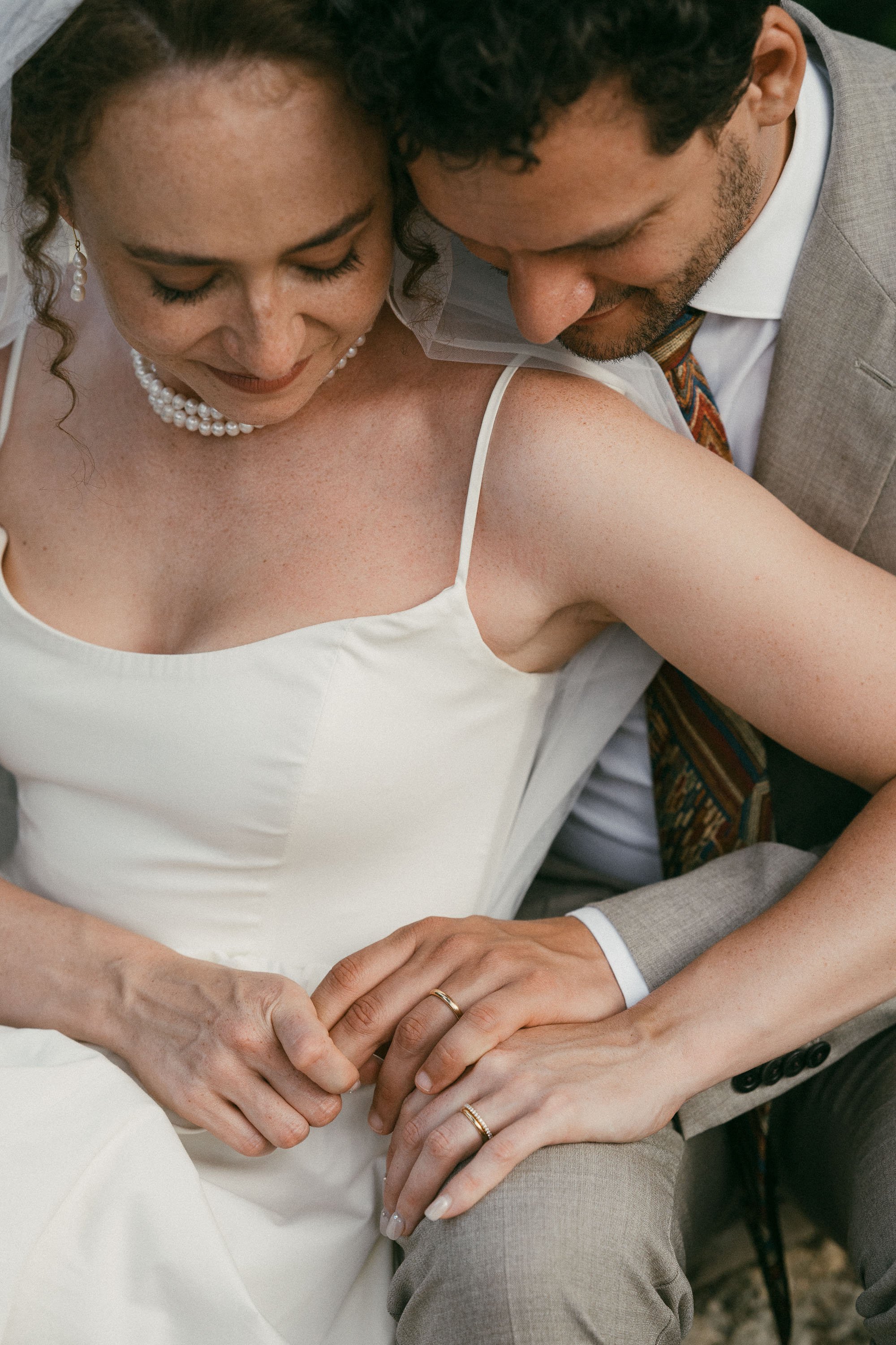 Close-up of a bride and groom holding hands, with their rings visible, during a wedding ceremony. The bride is wearing a white dress and pearl jewelry, and the groom is in a suit with a colorful tie.