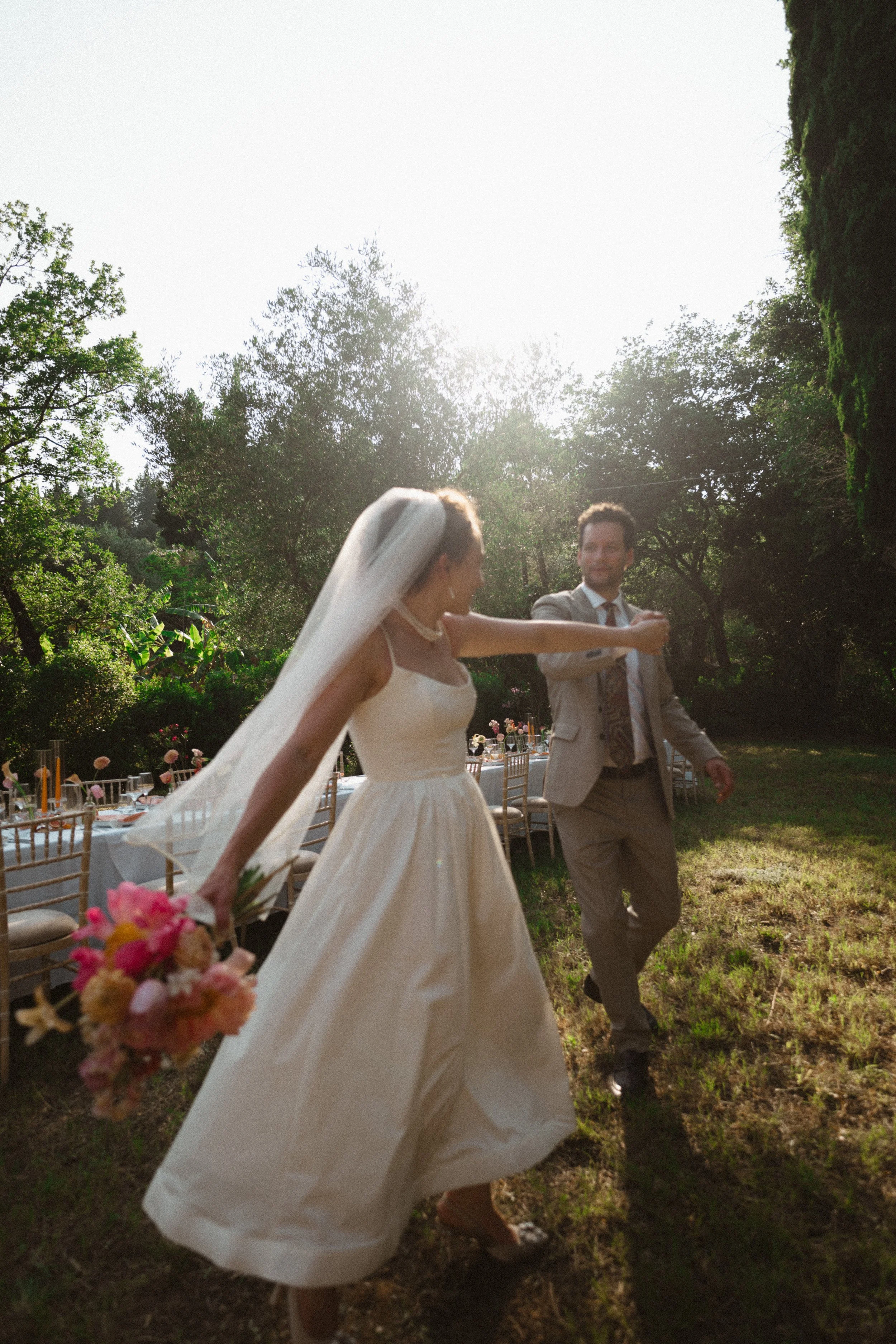 Bride and groom dancing outdoors at a wedding reception with tables and chairs in the background, lush greenery, and sunlight.