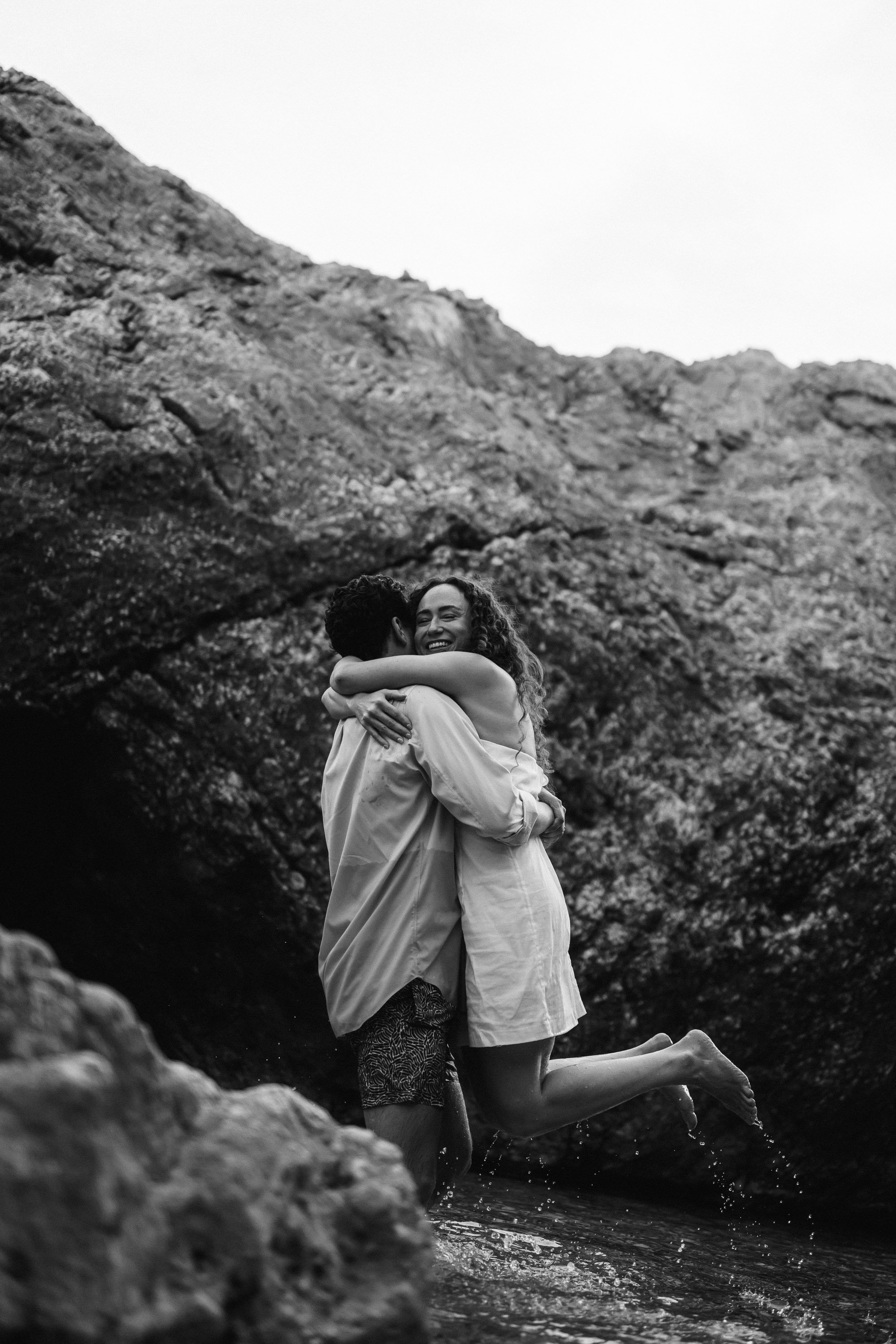 A couple embracing and smiling while jumping in a shallow stream in a rocky outdoor setting.