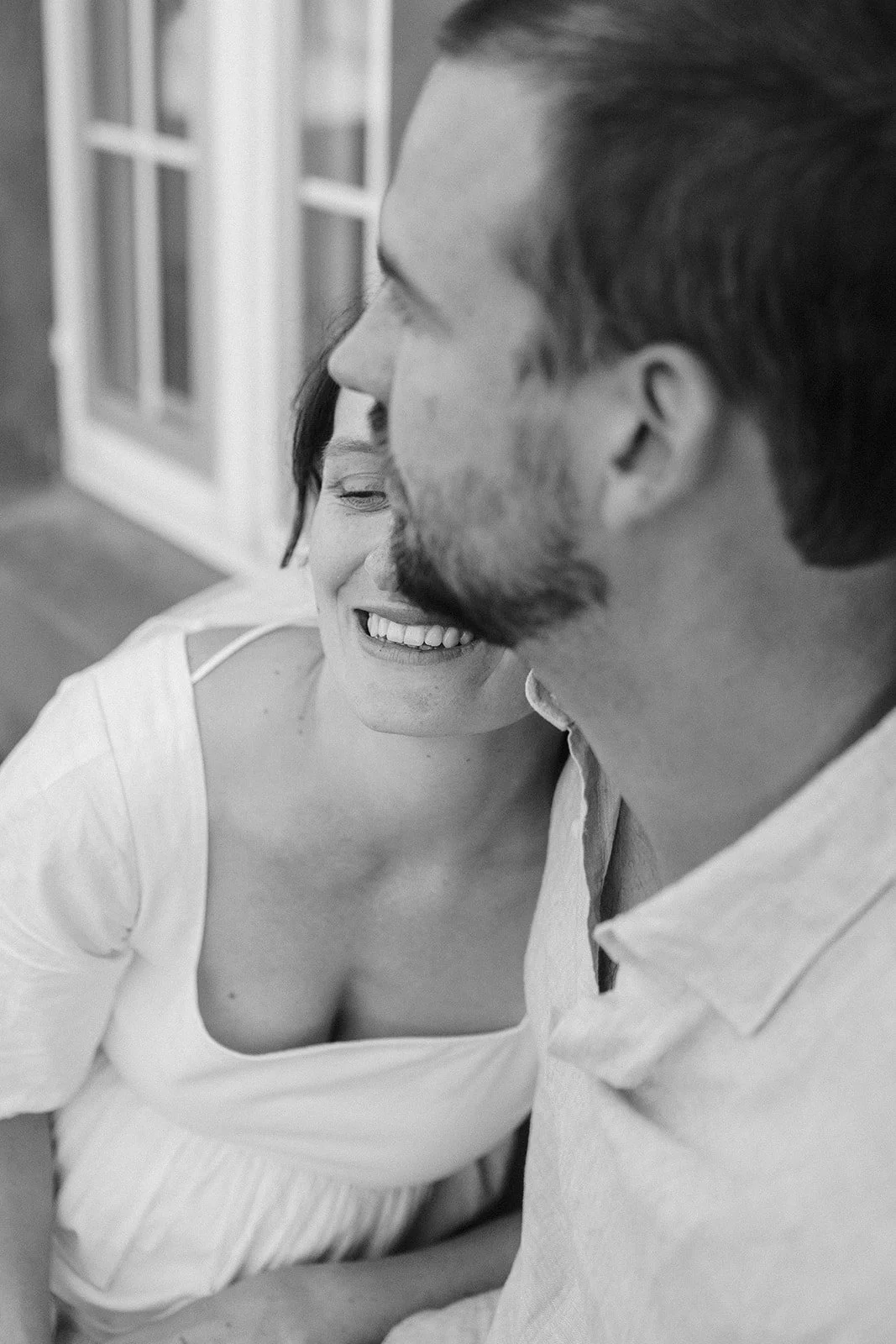 A black and white photo of a smiling woman and a man close together, with the woman's face partially hidden behind the man's face. They appear to be in a happy moment.