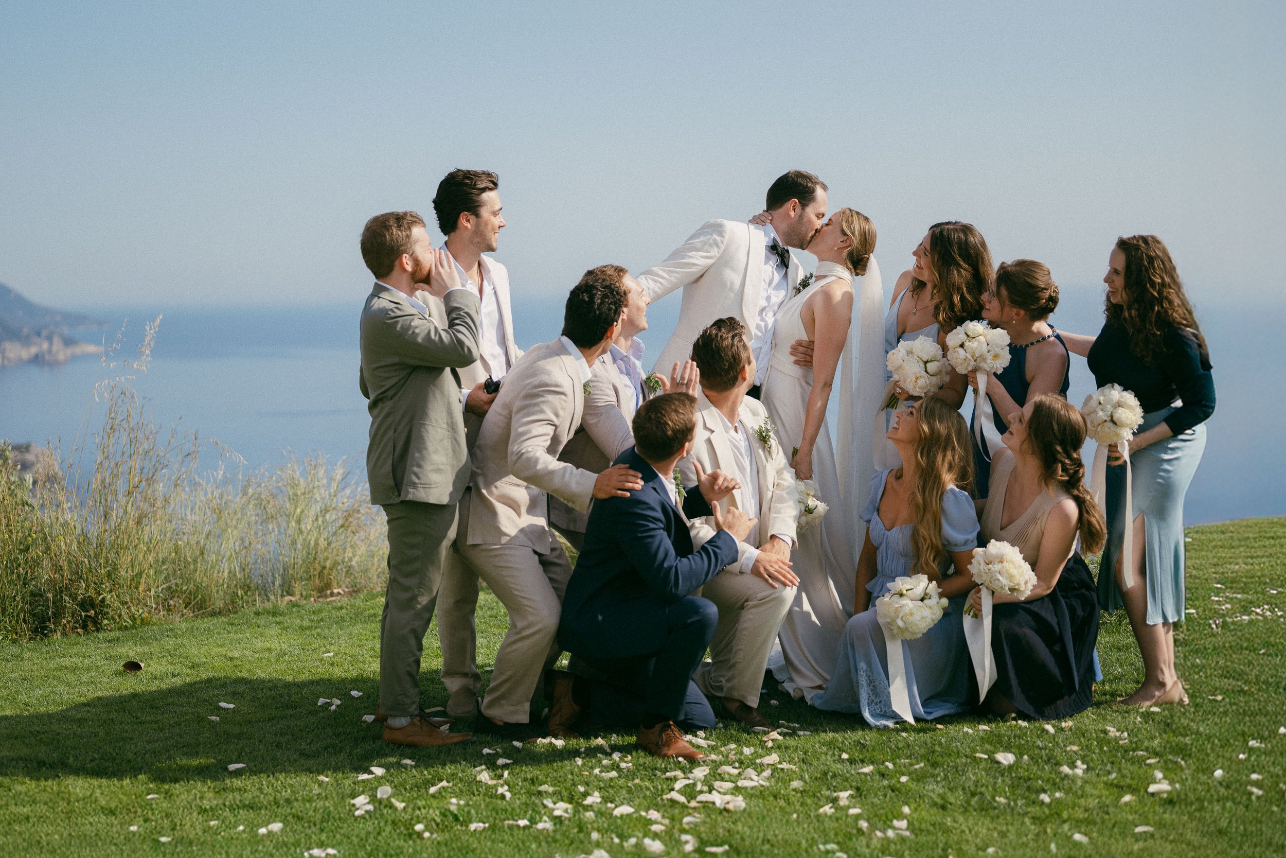 A wedding party outdoors with the ocean in the background, celebrating the newlywed couple who are kissing.