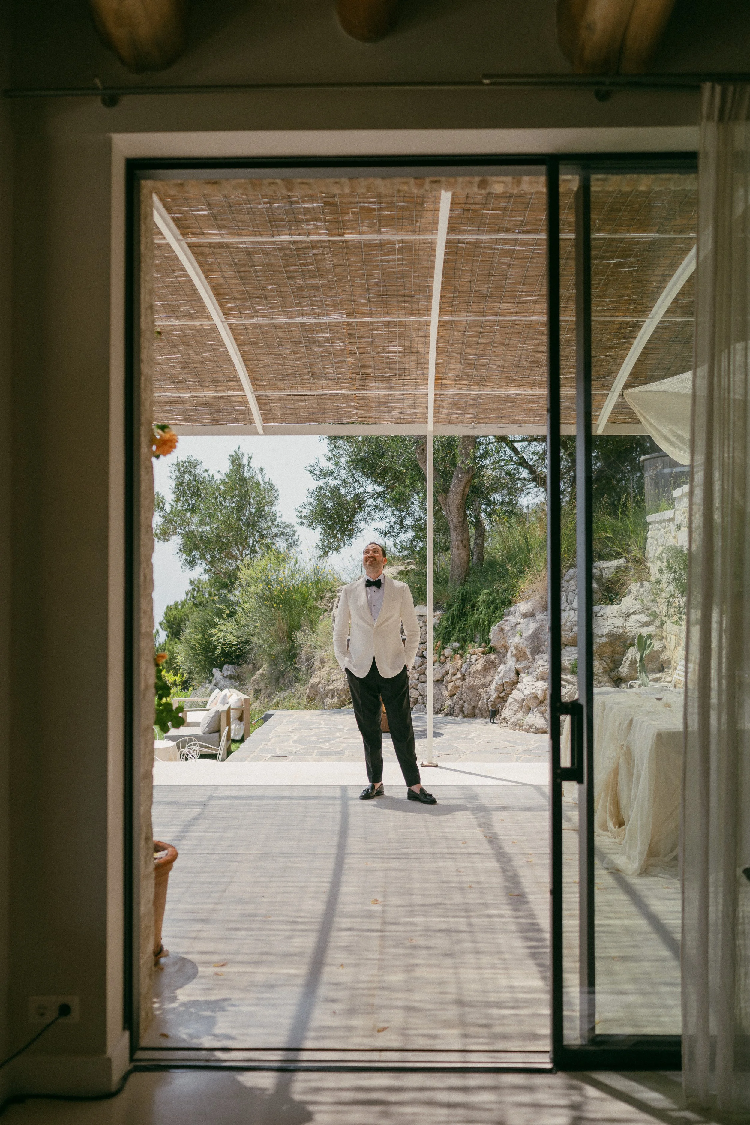 A man dressed in a tuxedo with a cream blazer, black bow tie, and black pants stands outside on a patio, viewed through a sliding glass door from inside a house. The patio has a wooden overhead cover and natural stone flooring, with trees and rocks i