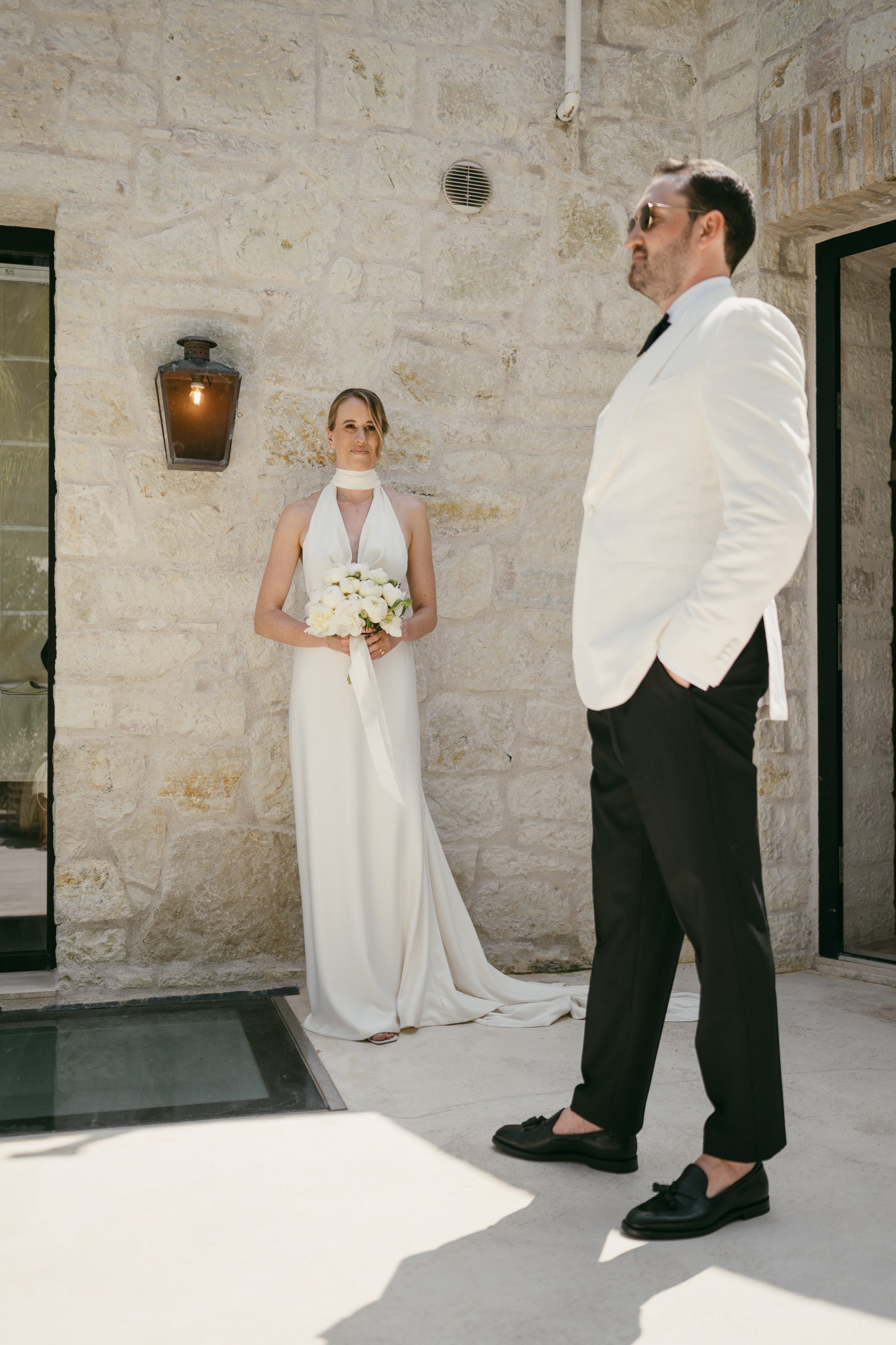 A bride in a white wedding dress holding a bouquet stands watching a groom in a white tuxedo jacket and black pants during an outdoor wedding ceremony against a stone wall.