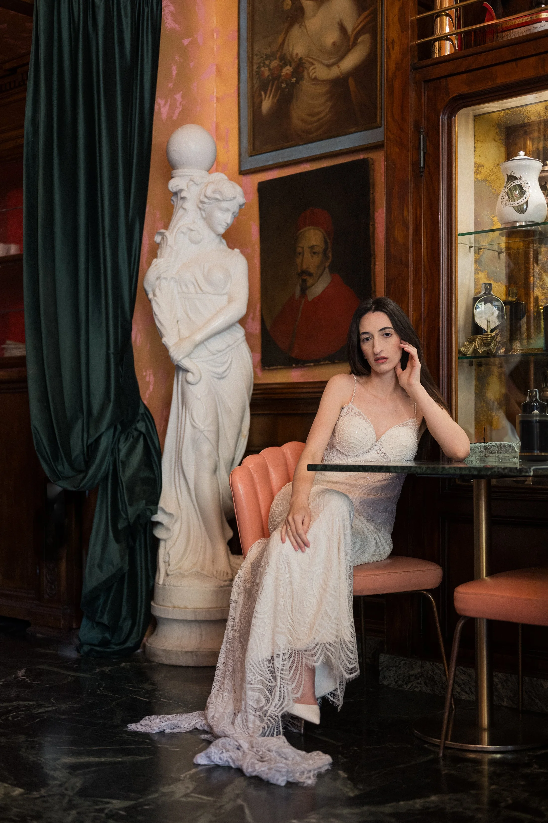 A woman in a white lace dress sitting at a table in an art-filled room with statues and portraits behind her.