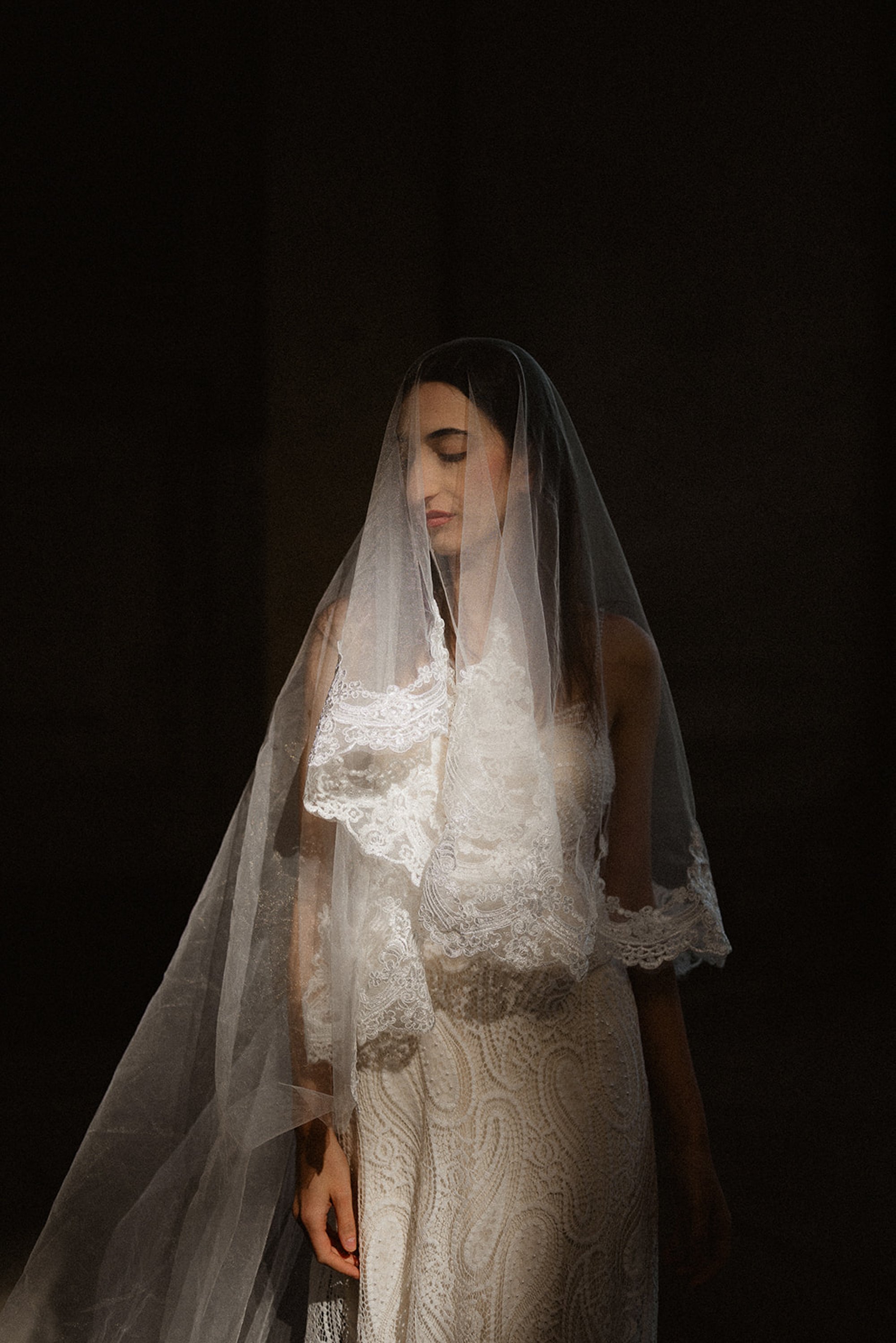 A woman in a lace wedding dress and veil, standing with her eyes closed, against a dark background.