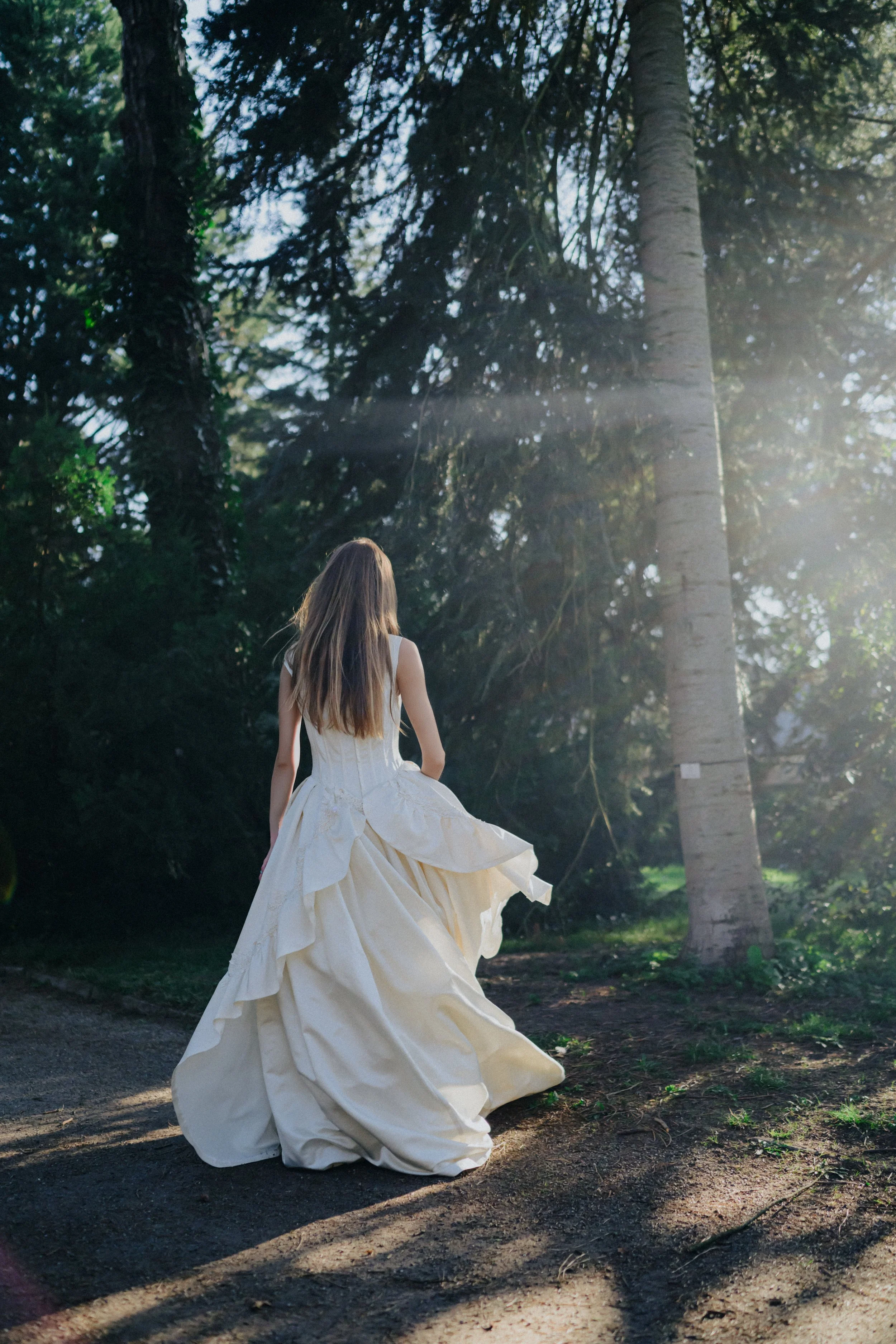 A woman in a flowing white dress walking through a forest with sunlight streaming through the trees.