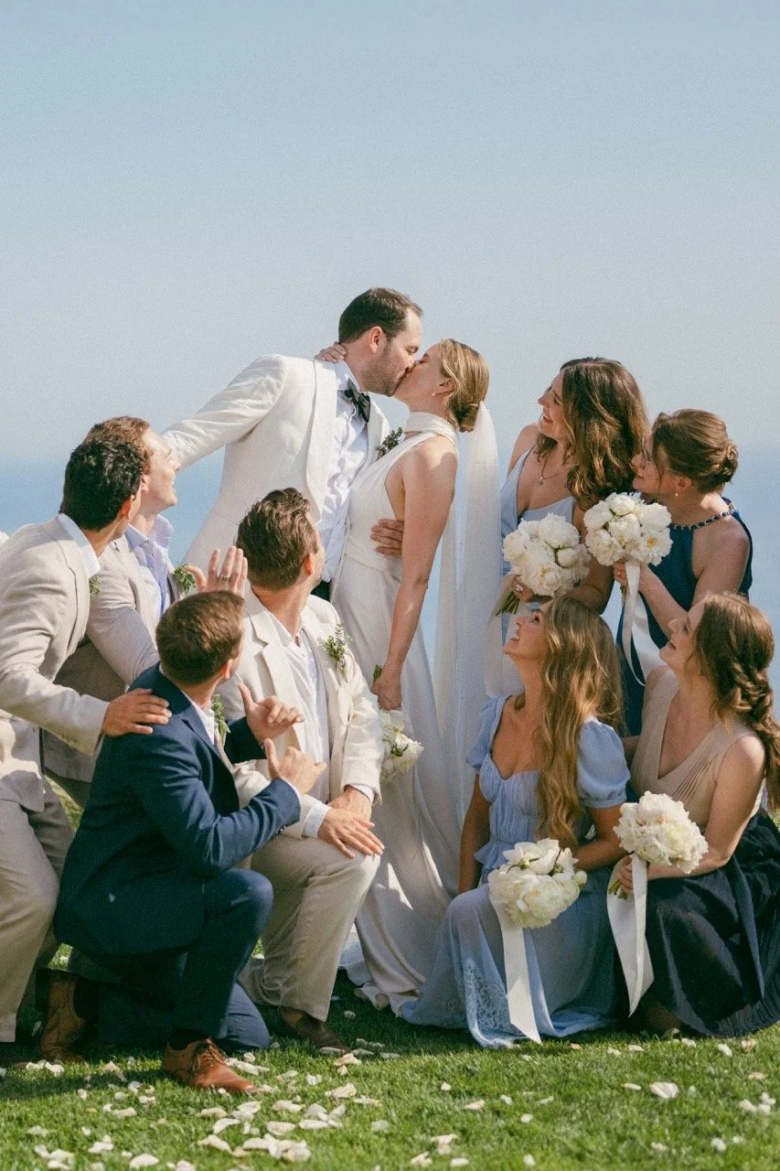 A wedding scene with a bride and groom sharing a kiss while surrounded by bridesmaids and groomsmen outdoors under a clear sky.