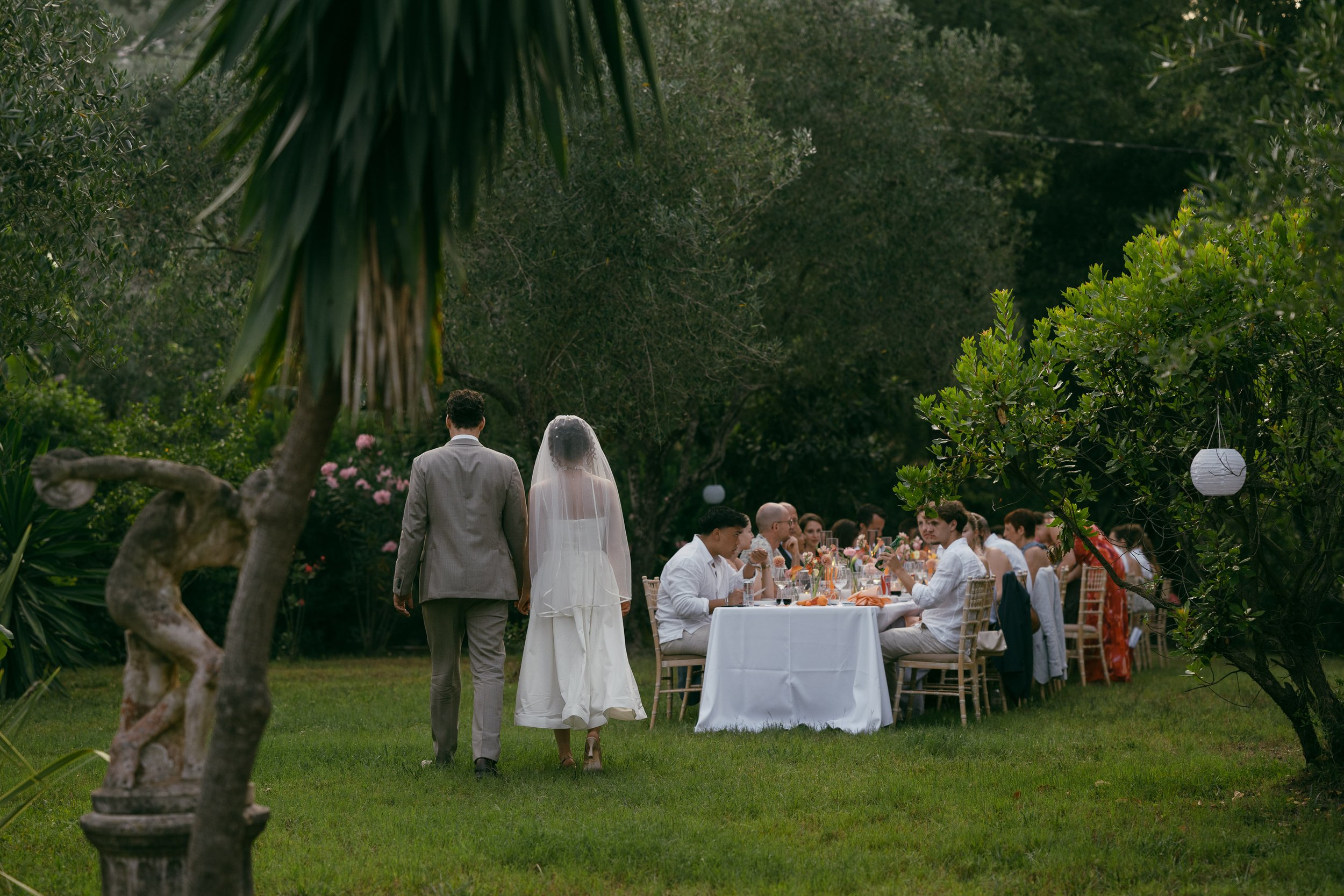 A wedding reception outdoors with a bride and groom walking towards a long table filled with guests. The scene is set in a lush garden surrounded by trees and greenery, with decorative lanterns hanging from the trees.