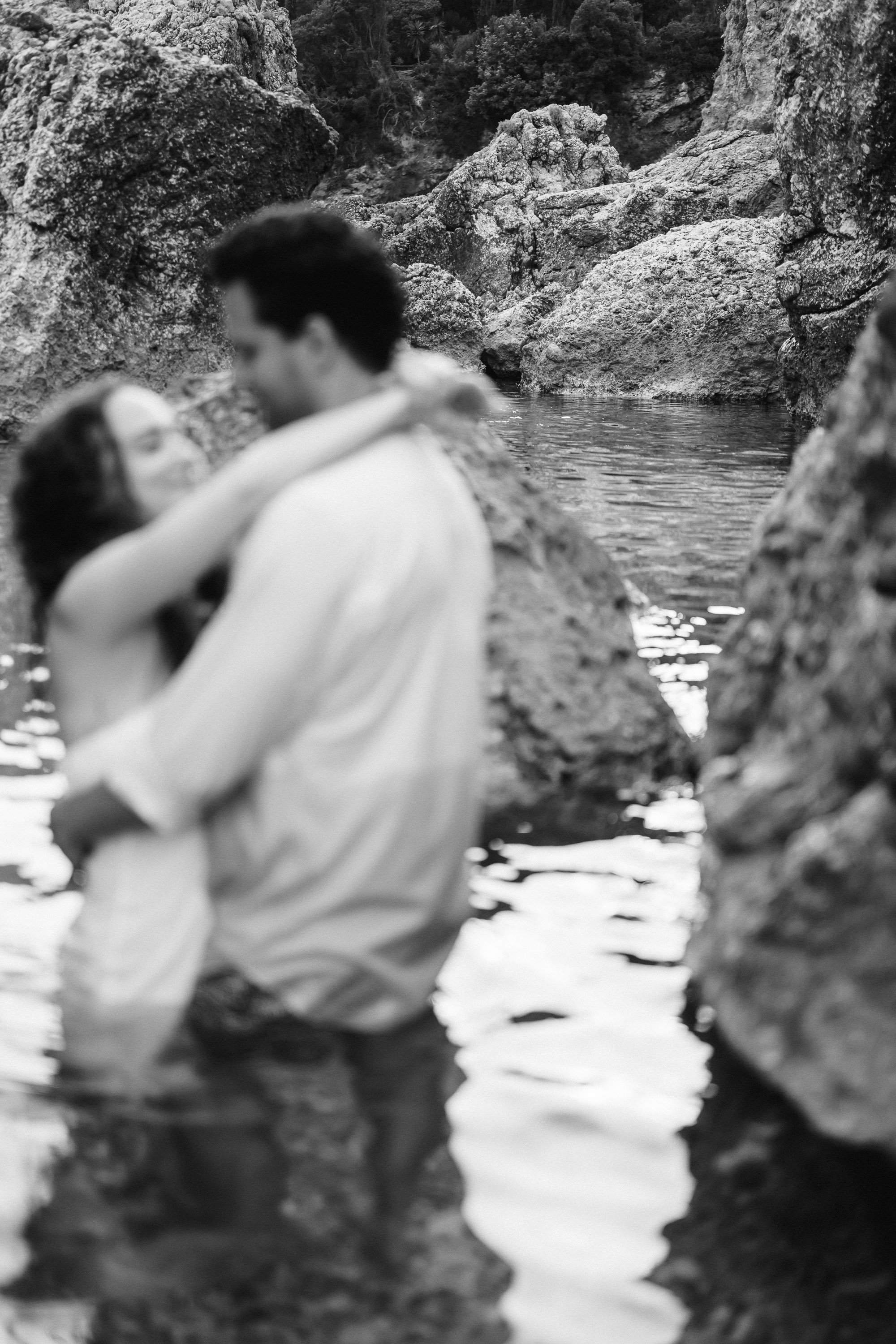 Black and white photo of a couple embracing in a rocky water setting, with rocks and water surrounding them.