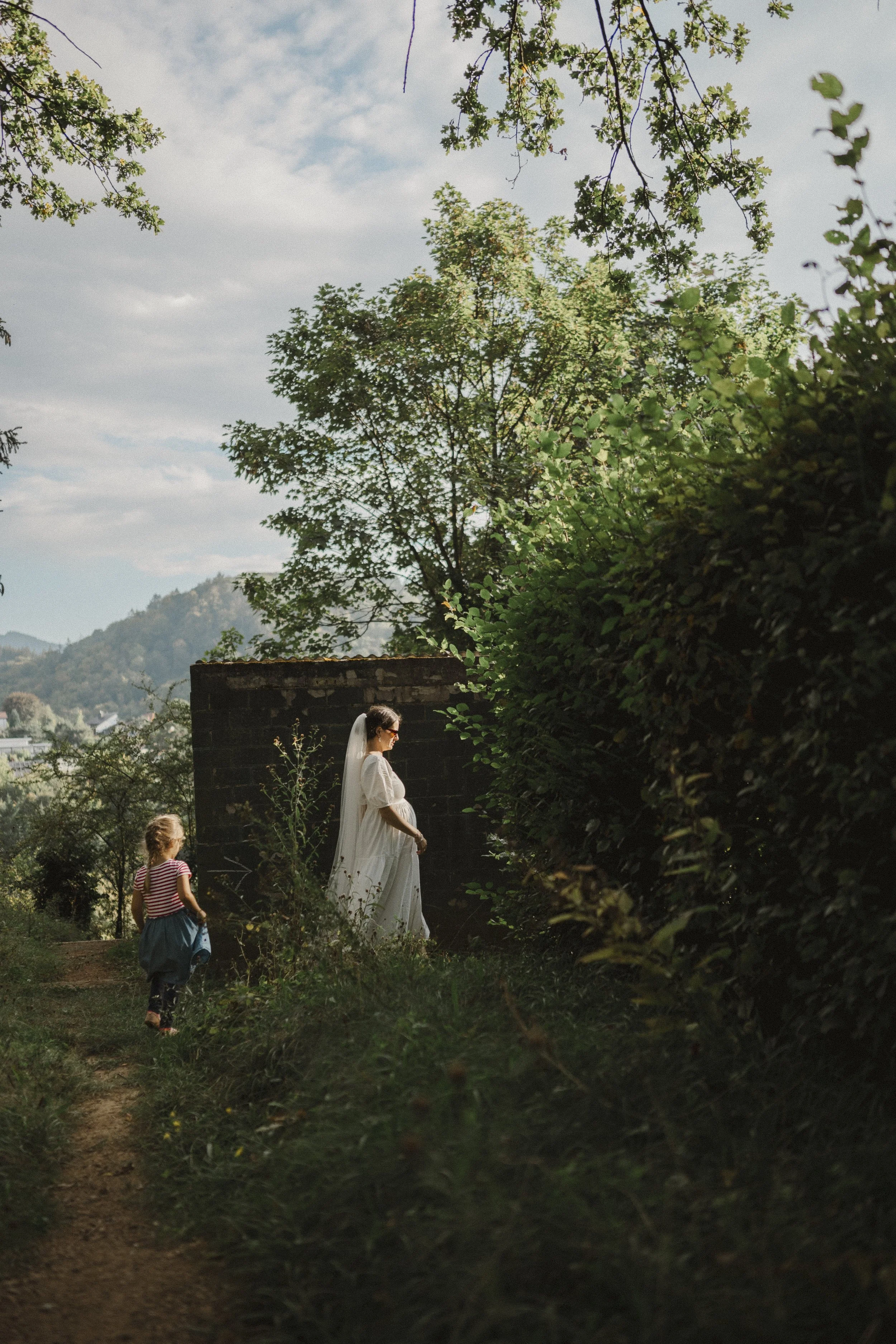A woman in a white dress, wearing a veil, standing on a grassy path next to a hedge and a stone wall, with a young girl walking behind her, on a sunny day with a partly cloudy sky and trees in the background.