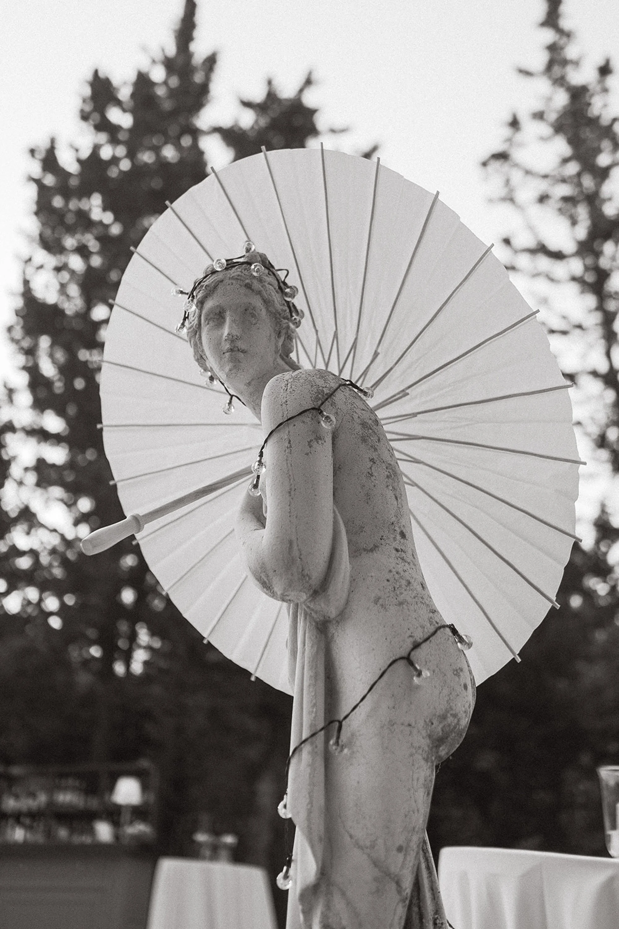 Black and white photo of an ancient statue of a woman with a parasol and decorative lights. The background shows trees.