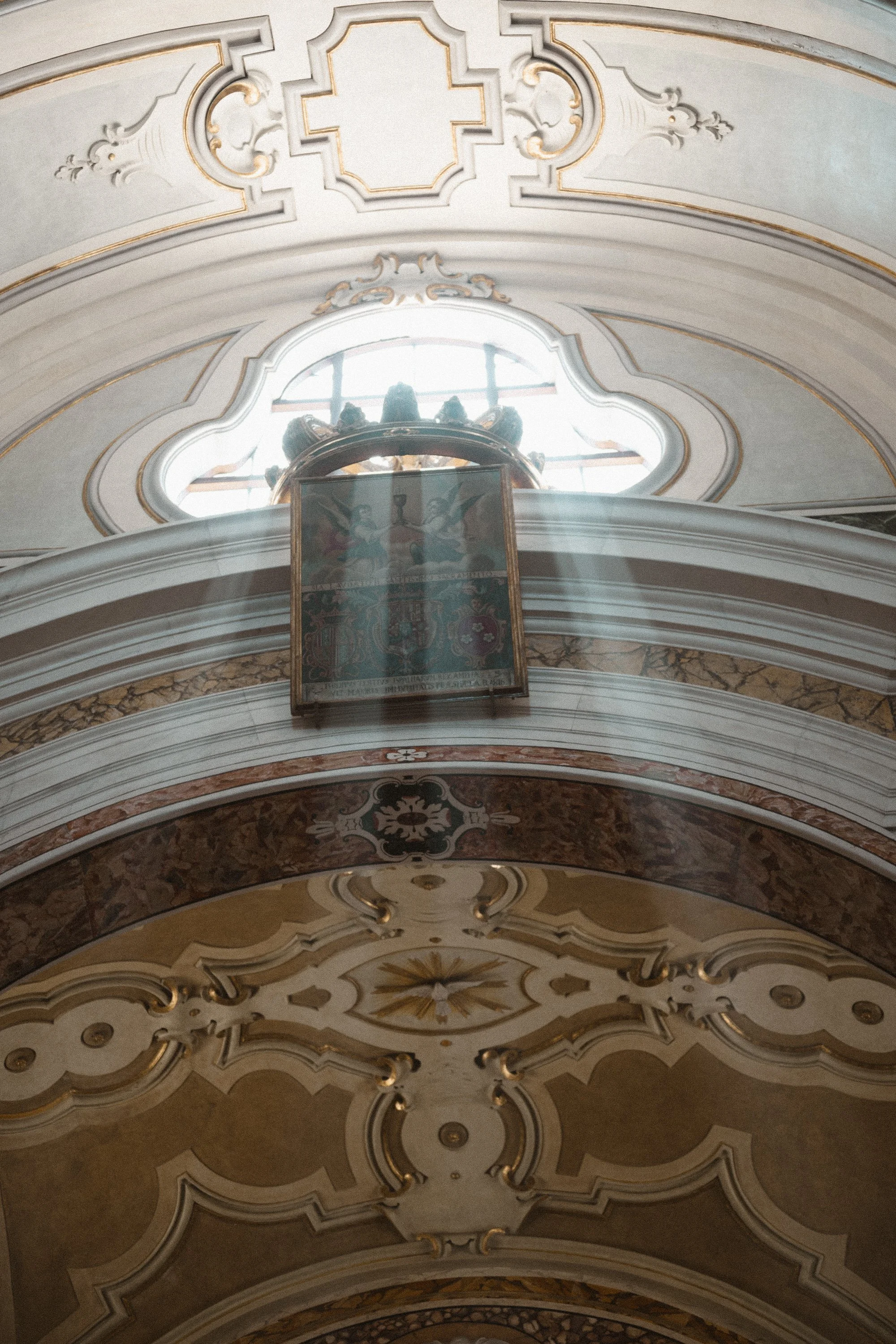 Interior view of a church ceiling with ornate moldings, a window with sunlight rays, and a religious painting or icon.